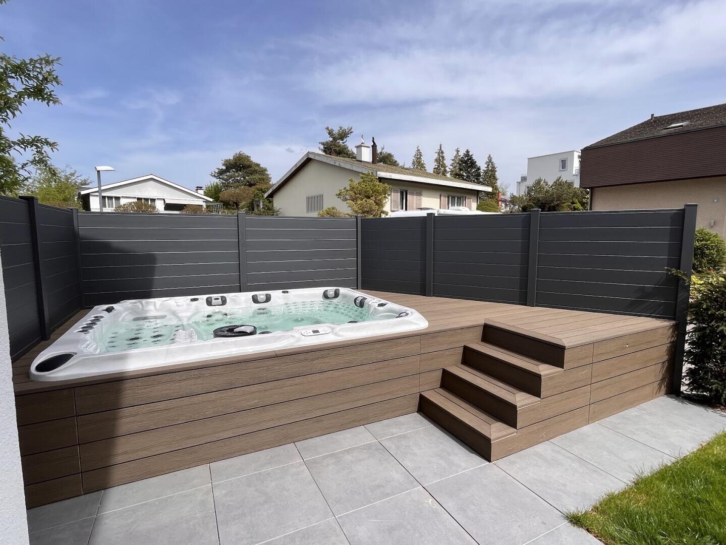 Ein moderner Whirlpool mit sprudelndem Wasser steht an einem sonnigen Tag auf einer Holzterrasse mit Stufen, umgeben von einem grauen Sichtschutzzaun in einem privaten Hinterhof.