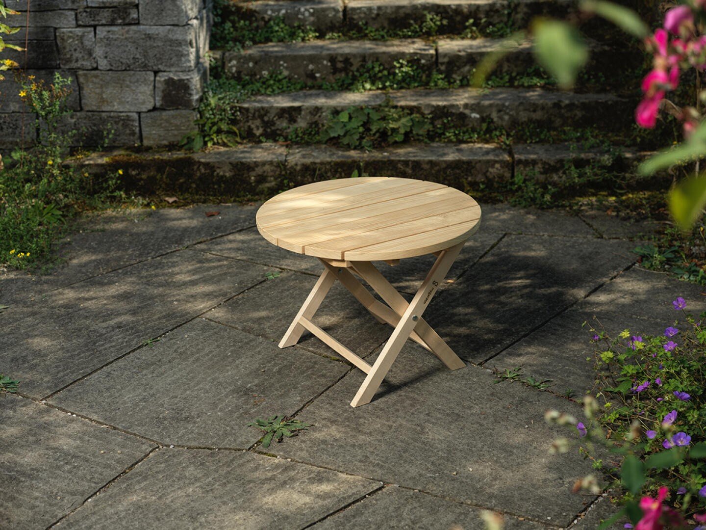 Ein kleiner runder Klapptisch aus Holz steht auf einer Steinterrasse mit Steinstufen und Grünpflanzen im Hintergrund, umgeben von einigen blühenden Pflanzen.