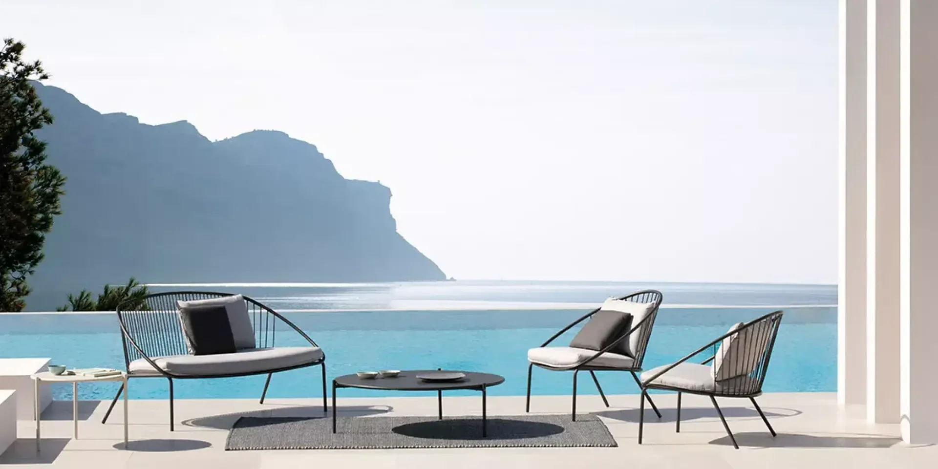 Moderne Außenterrasse mit minimalistischen Möbeln mit Blick auf einen Infinity-Pool und das ruhige Meer, mit einem Berg und einem klaren Himmel im Hintergrund. Die Szene ist heiter und stilvoll.