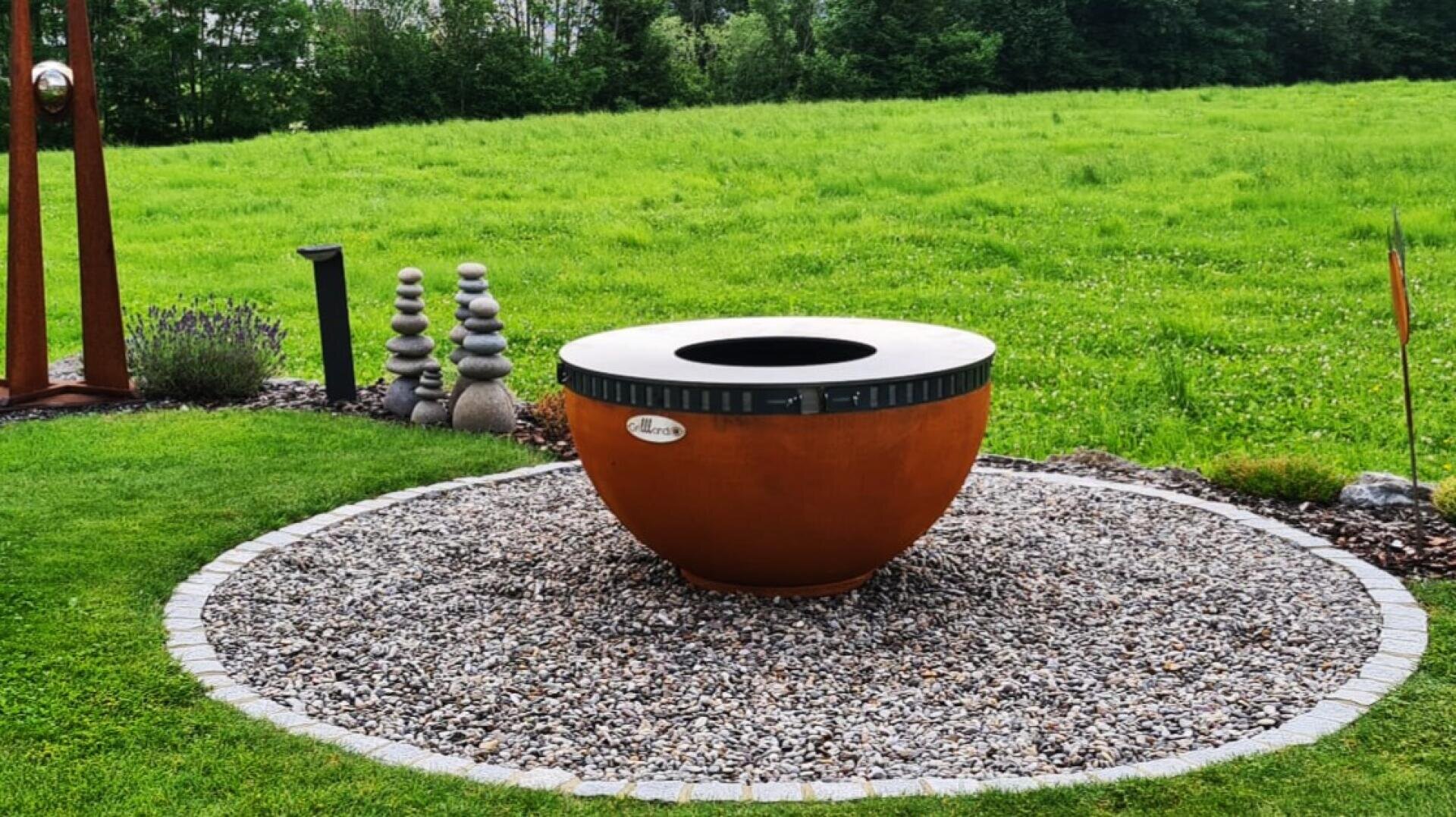 Eine große, runde, rostfarbene Feuerstelle befindet sich auf einem mit Ziegelsteinen eingefassten Kieskreis, umgeben von grünem Gras. Im Hintergrund befinden sich gestapelte Steinskulpturen und Bäume.