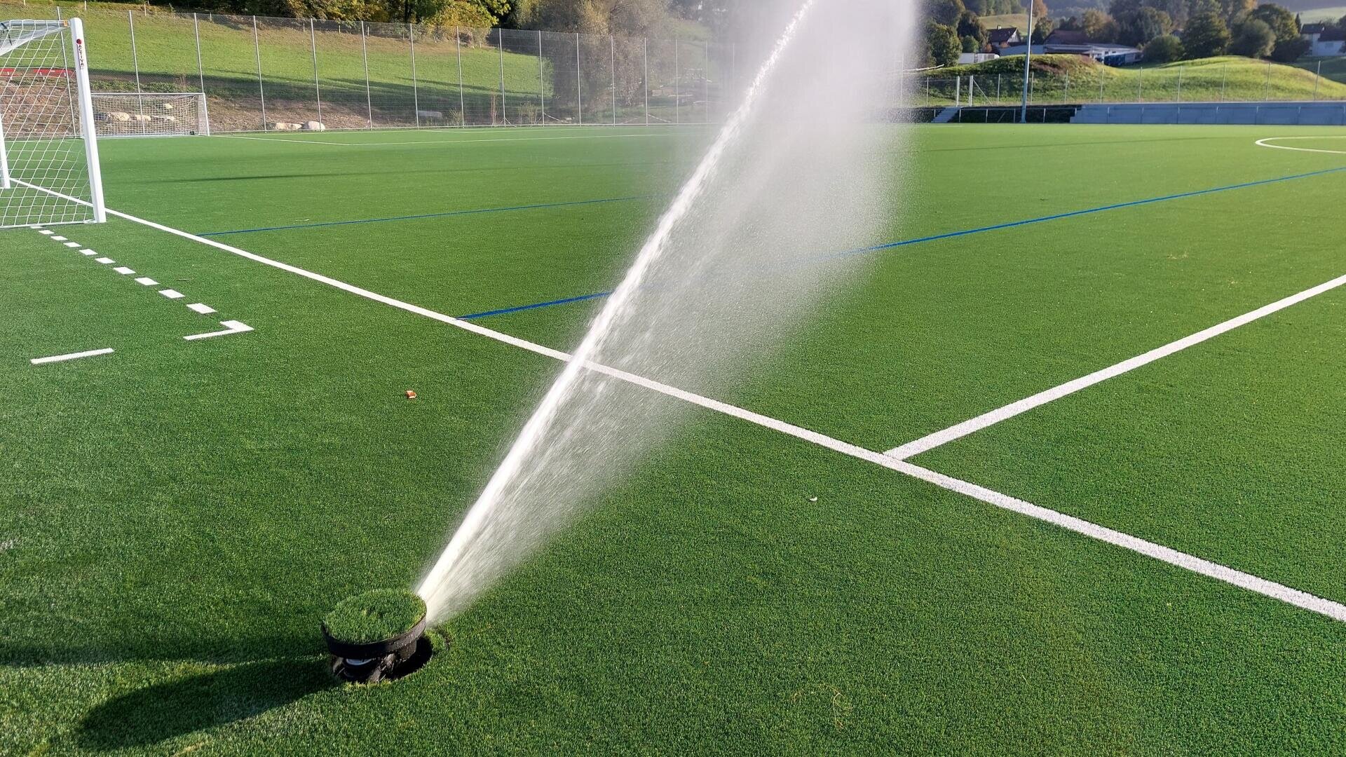 A sprinkler sprays water onto a green artificial turf soccer field near the corner and goal lines on a sunny day. White field markings and a goalpost are visible in the background.