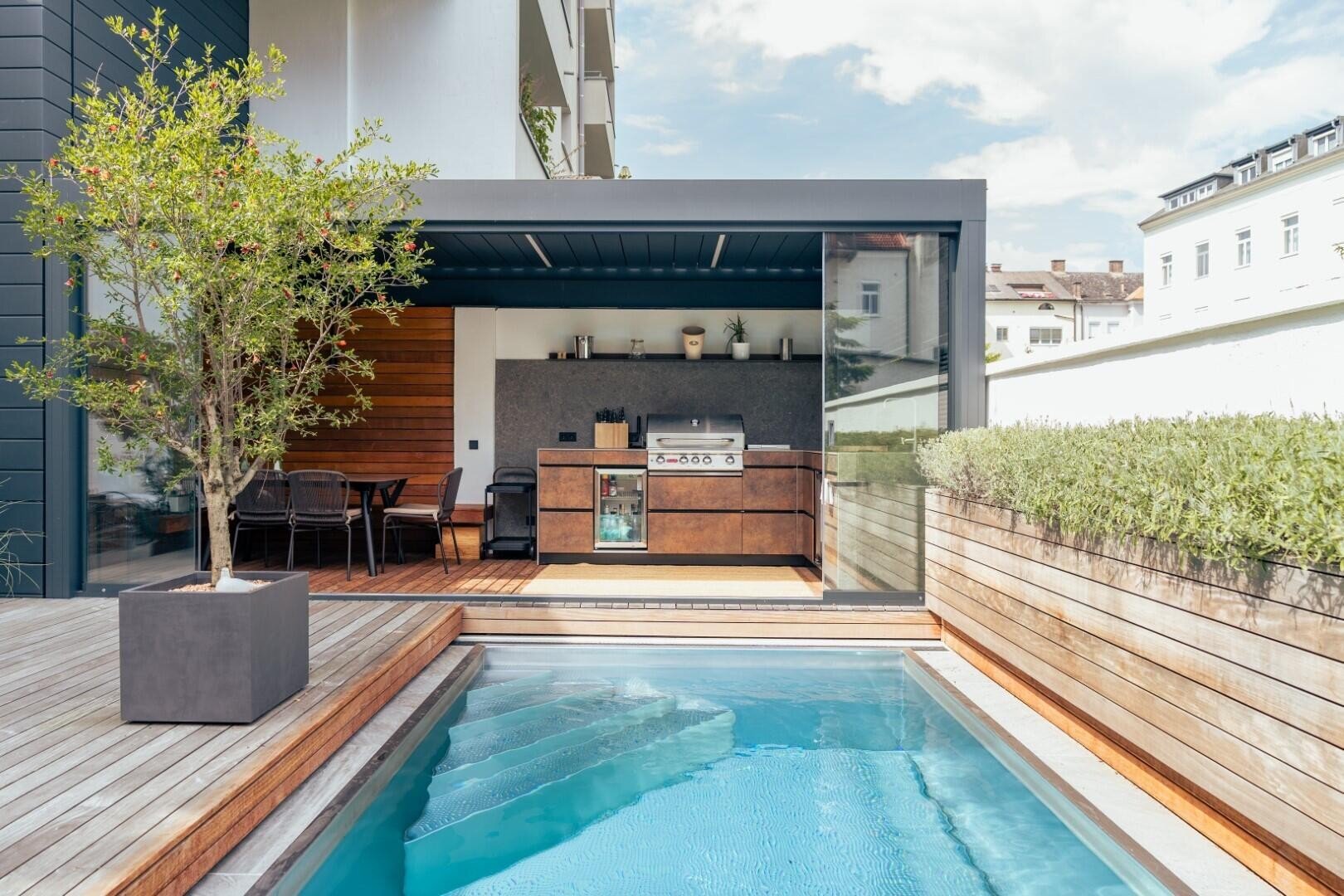 Moderner Garten mit einem kleinen rechteckigen Pool, Holzdeck, Topfbaum und einer Außenküche mit Grill und Essbereich unter einer überdachten Terrasse. Hohe Hecke Linien auf der rechten Seite für die Privatsphäre.