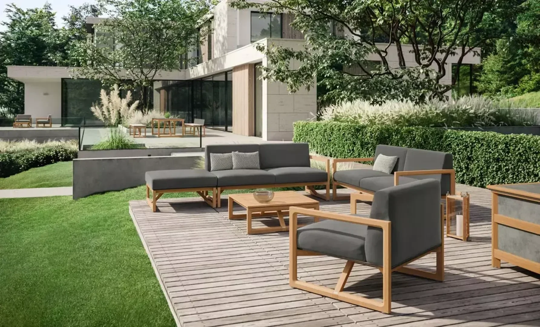 Modern outdoor patio with wooden deck, gray cushioned seating, and a coffee table, surrounded by lush greenery, hedges, and a contemporary house in the background.