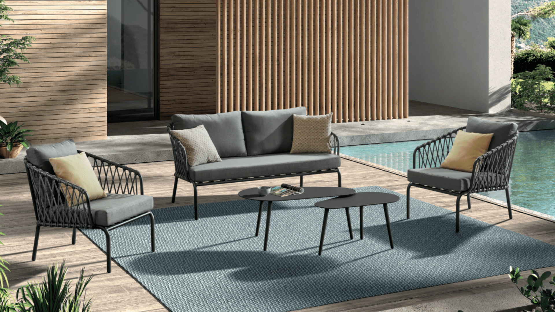 Terrassenset mit einem grauen Sofa, zwei Sesseln mit gelben Kissen, zwei schwarzen Couchtischen und einem blauen Teppich auf einer Holzterrasse neben einem Pool. Im Hintergrund sind ein modernes Gebäude und Grünflächen zu sehen.