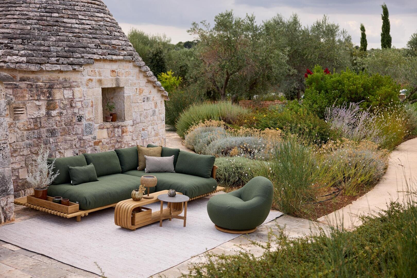 Ein Sitzbereich im Freien mit einem grün gepolsterten Sofa und Sessel, einem kleinen Couchtisch und einem Teppich befindet sich neben einem rustikalen Steinhaus, umgeben von üppigem Grün und Gartenwegen.