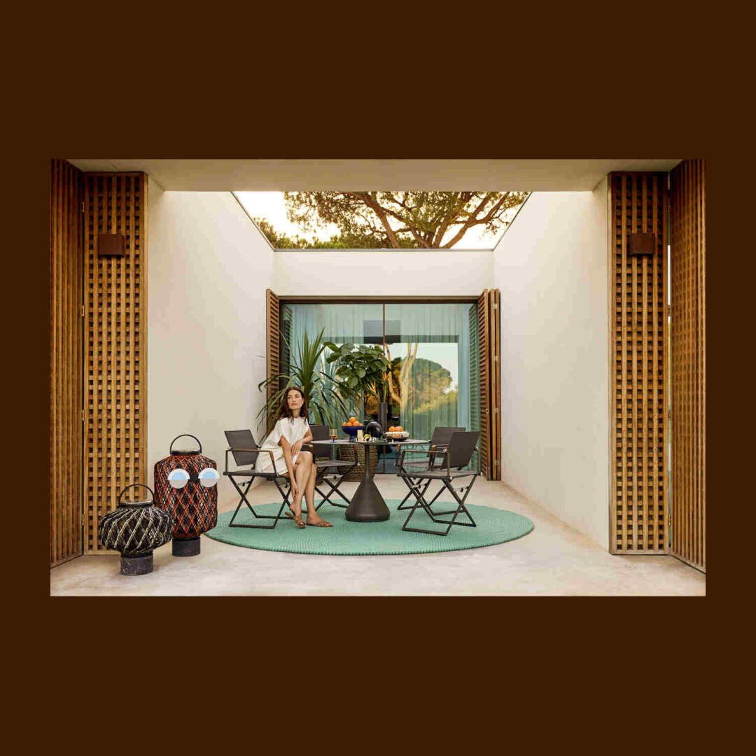 Eine Frau sitzt auf einem Stuhl an einem Essplatz im Freien, umgeben von Pflanzen und Dekoration, auf einem runden grünen Teppich. Der Bereich ist von hohen hölzernen Paravents und Glastüren umgeben, durch die das Sonnenlicht einfällt.