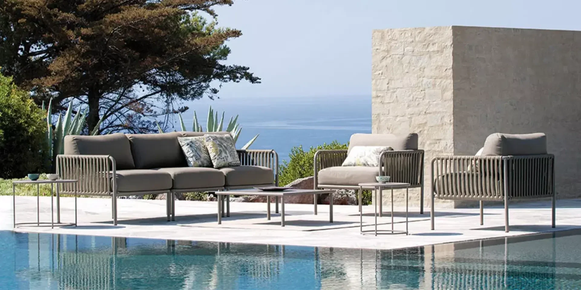 Moderne Terrassenmöbel, darunter ein Sofa, Stühle und Tische, sind auf einer Steinterrasse neben dem Swimmingpool aufgestellt. Der Blick geht auf das Meer, mit Bäumen und einer Steinmauer im Hintergrund.