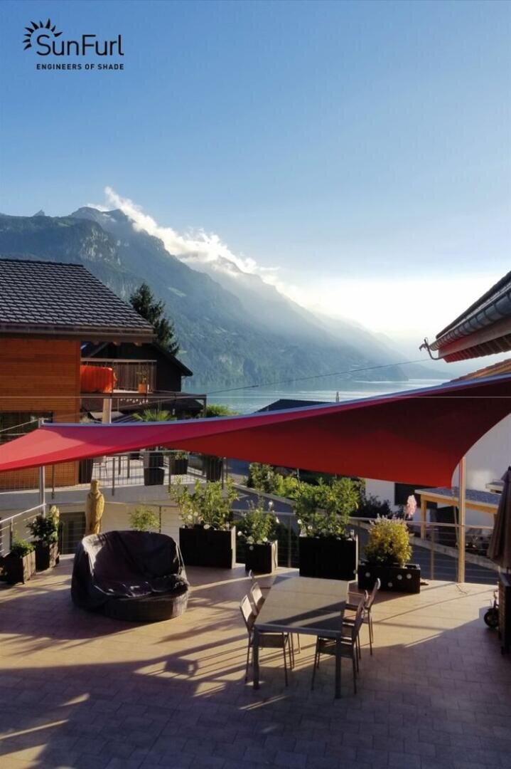 Eine Terrasse mit roten Sonnensegeln, Tisch und Stühlen, umgeben von Topfpflanzen, mit Blick auf einen See und Berge unter einem klaren Himmel; SunFurl Engineers of Shade-Logo in der oberen linken Ecke.
