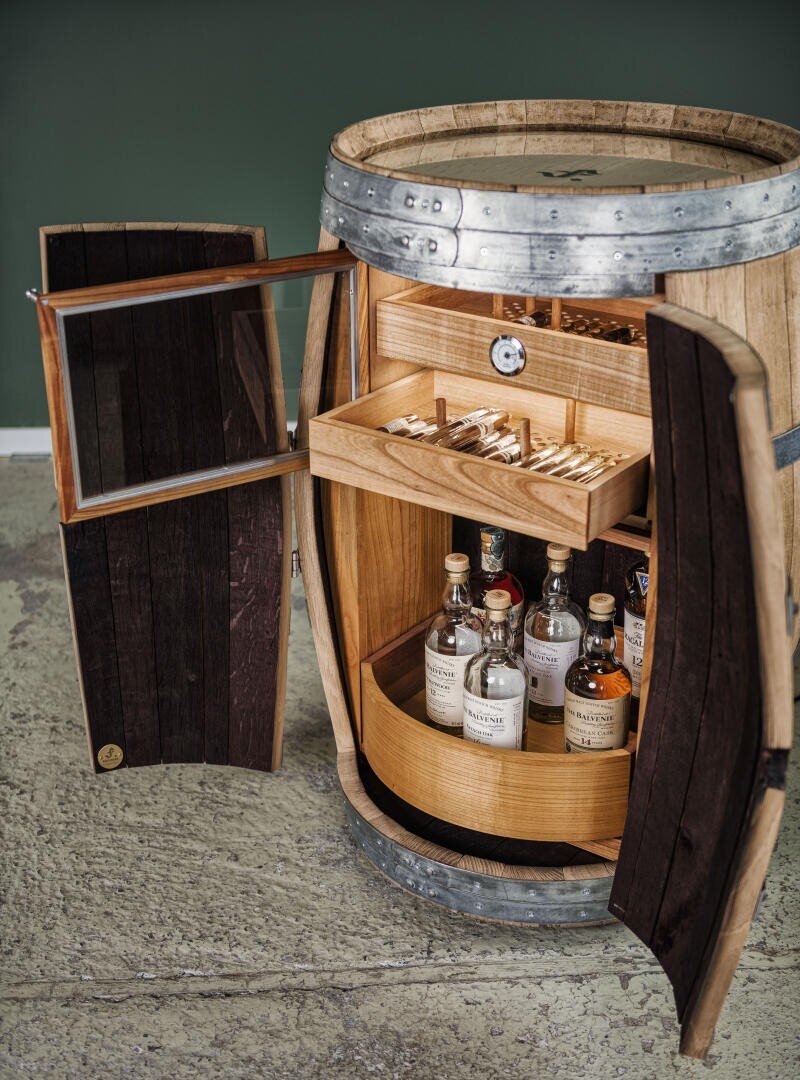 The front of a wooden barrel with metal straps is open, revealing shelves with bottles of schnapps on the floor and cigars in a drawer above, set against a green wall and a concrete floor.