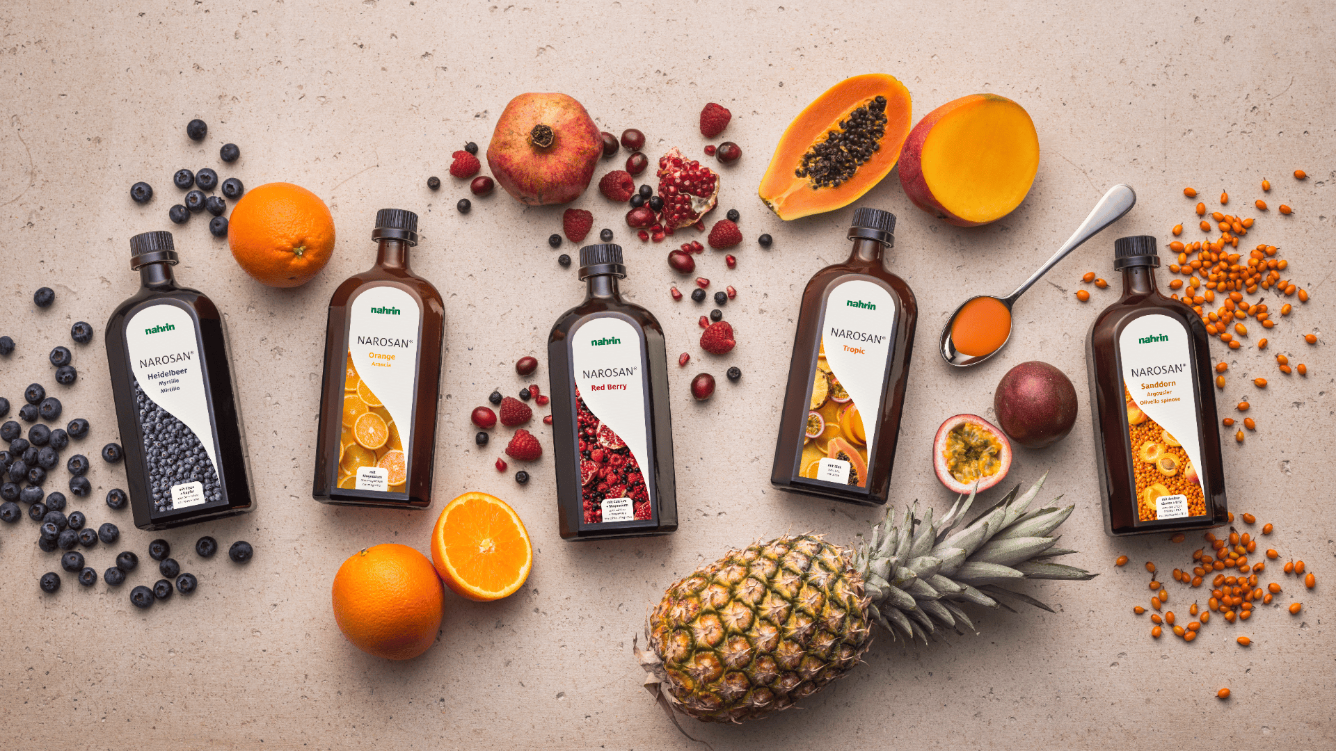 Fünf braune Flaschen mit Fruchtkonzentrat stehen auf einer hellen Fläche, umgeben von frischen Früchten wie Orangen, Ananas, Blaubeeren, Papaya, Granatäpfeln und Beeren, daneben ein Löffel mit Orangenflüssigkeit.