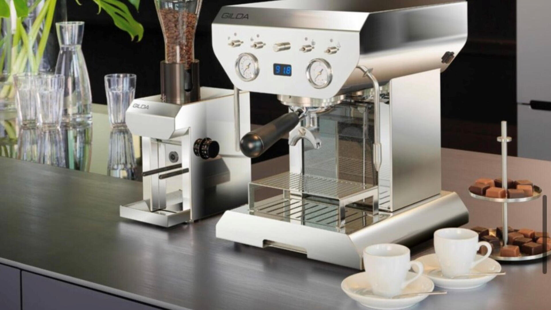 A modern stainless steel espresso machine and coffee grinder are on a kitchen counter next to two white cups with saucers, a plate of chocolates, and glassware in the background.
