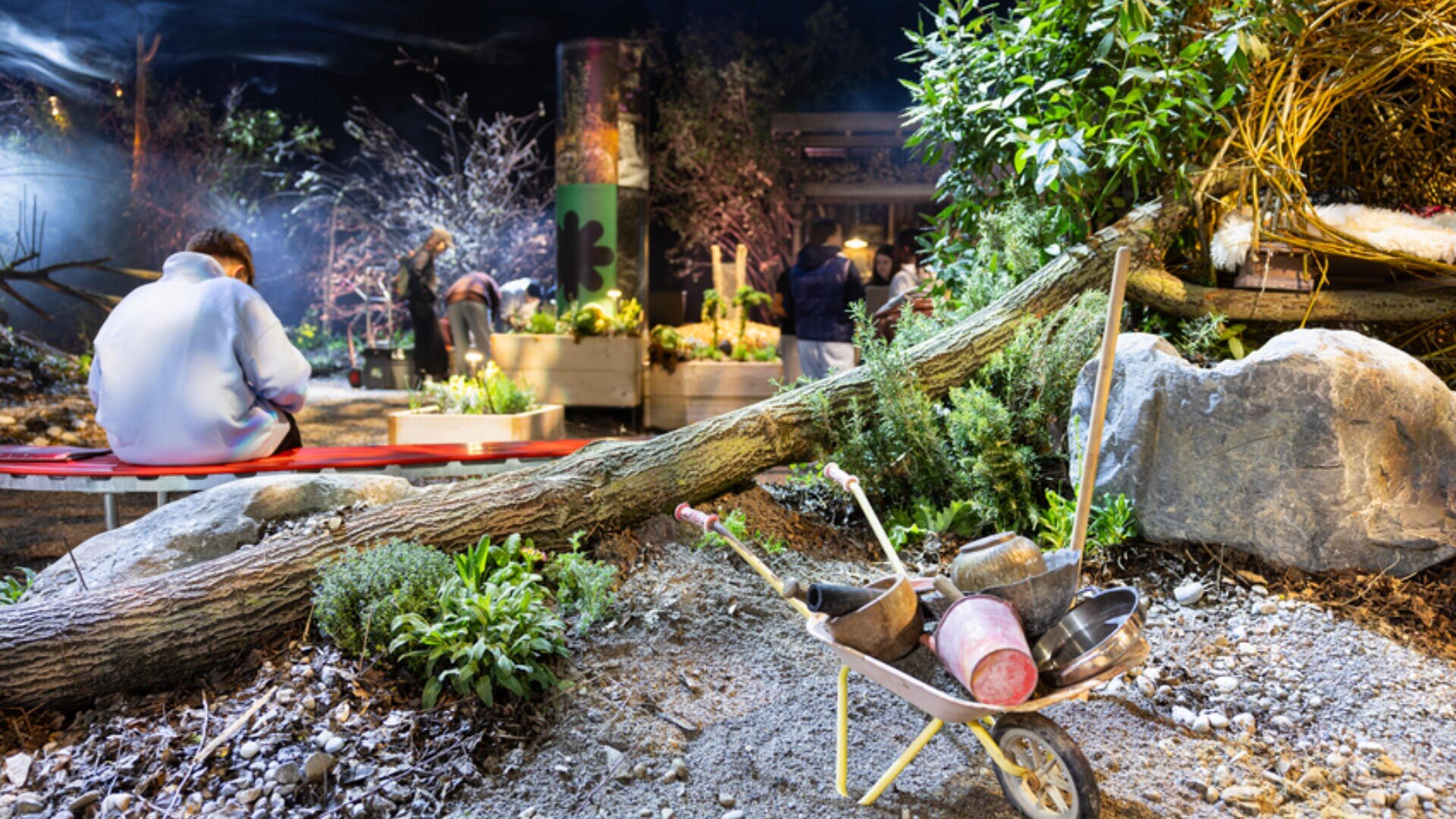 Eine nächtliche Gartenszene im Freien mit Menschen, die sich in der Nähe von Hochbeeten versammelt haben. Im Vordergrund sind ein umgestürzter Baum, Felsen und eine mit Töpfen und Gartengeräten gefüllte Schubkarre zu sehen. Der Bereich ist mit weichem Licht beleuchtet.