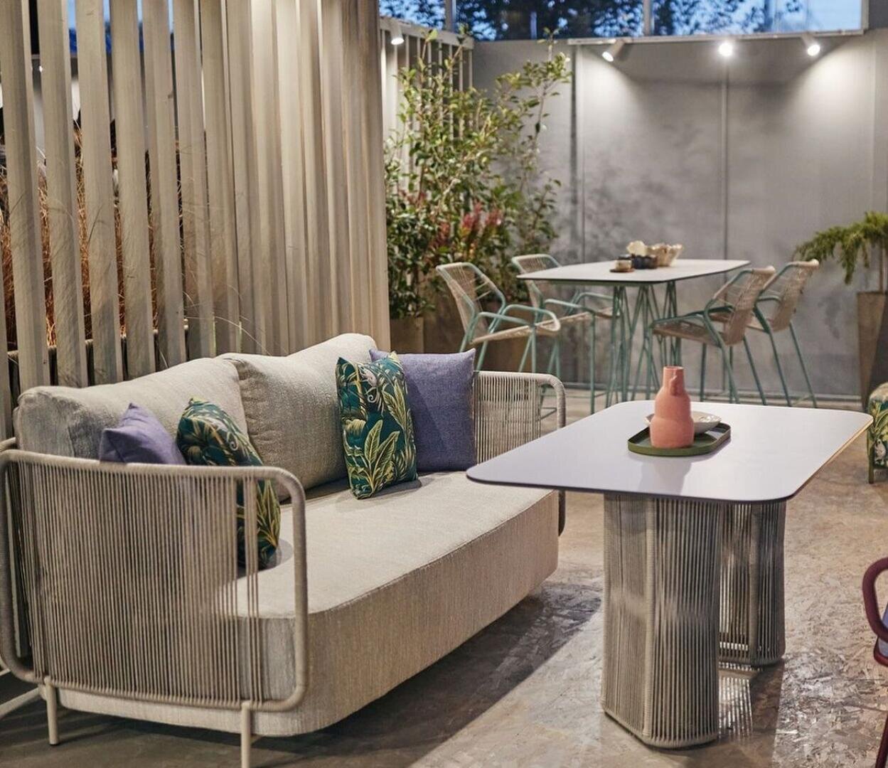 Ein moderner Sitzbereich im Freien mit einem hellen Sofa, dekorativen Kissen, einem eleganten Couchtisch mit einer rosafarbenen Vase und einem Esstisch mit Stühlen im Hintergrund, umgeben von Pflanzen und sanfter Beleuchtung.