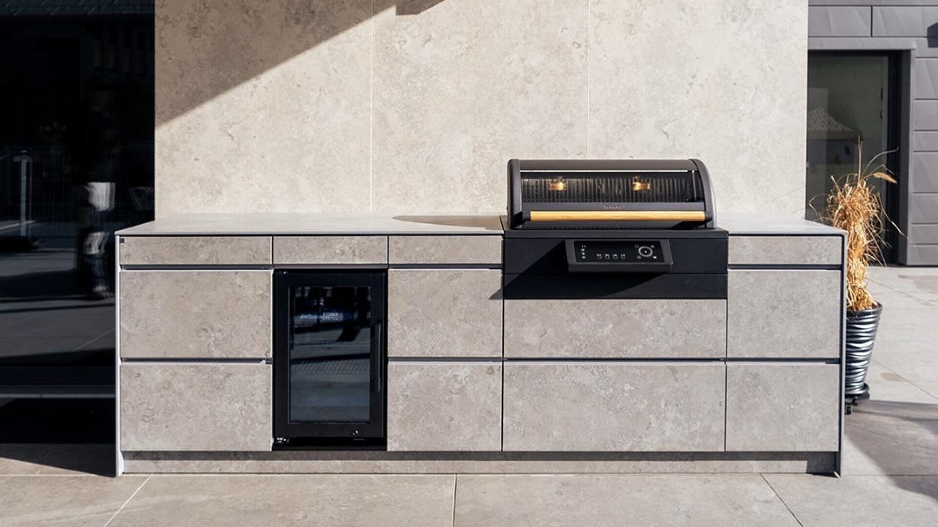 A modern outdoor kitchen with cabinets made of light-colored stone, a built-in mini fridge with glass door and an elegant grill with open lid and two lamps inside, which stands on a stone terrace.