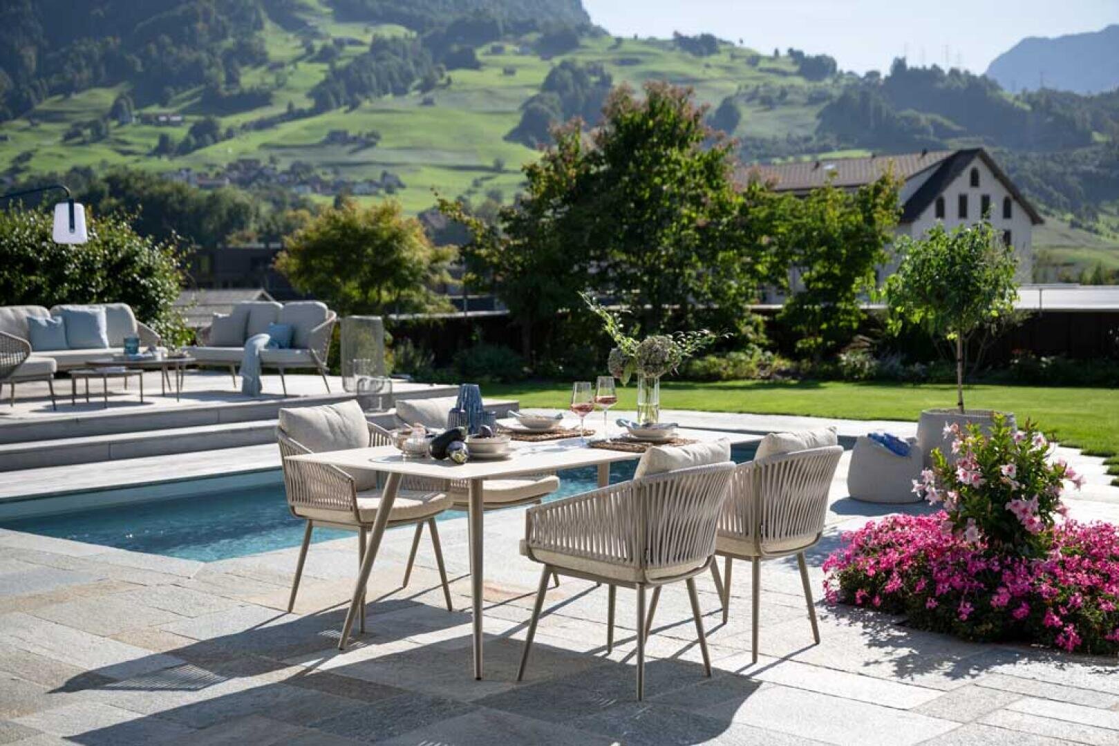 Ein moderner Esstisch mit vier Stühlen steht neben dem Swimmingpool, umgeben von üppigem Grün, blühenden Blumen und dem Blick auf Berge und traditionelle Häuser im Hintergrund an einem sonnigen Tag.