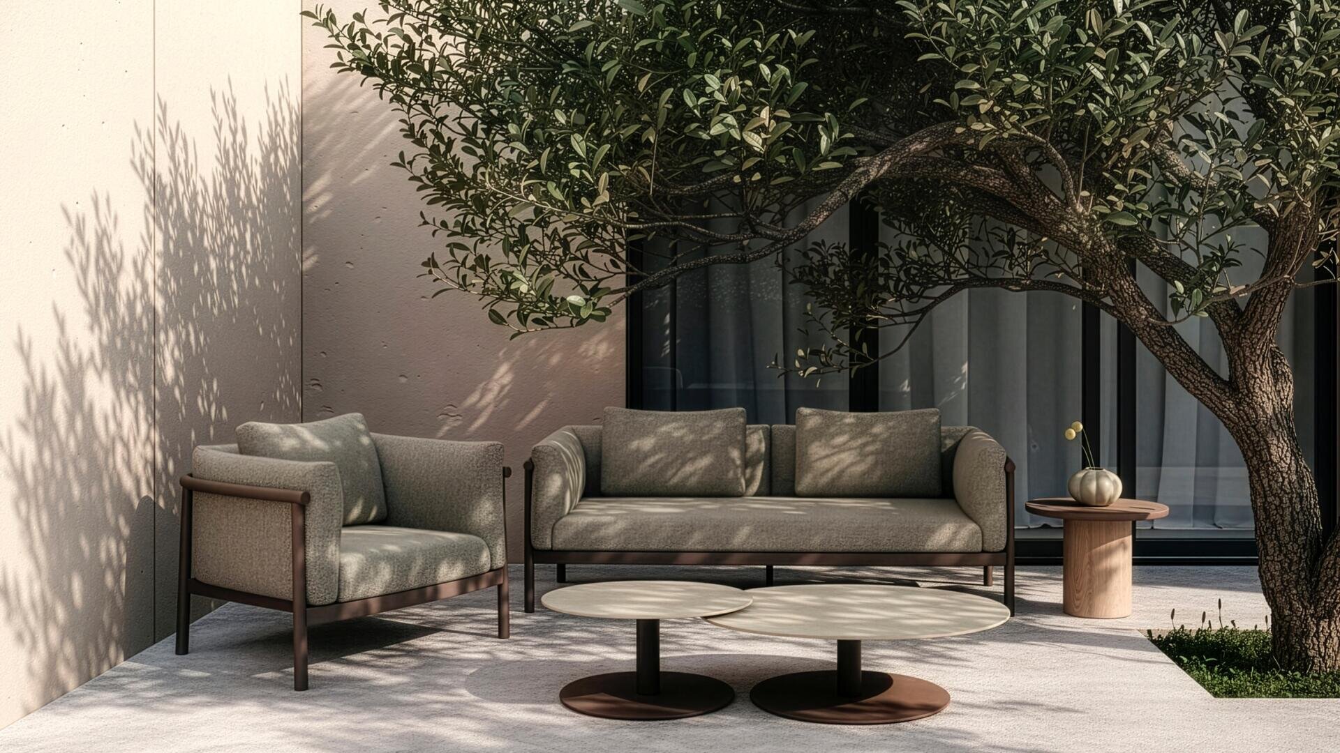 Eine moderne Außenterrasse mit einem gepolsterten Sessel, einem Sofa, zwei runden Couchtischen und einer kleinen Vase auf einem Beistelltisch, die von einem Olivenbaum beschattet und von einer beigen Wand und Glastüren begrenzt wird.