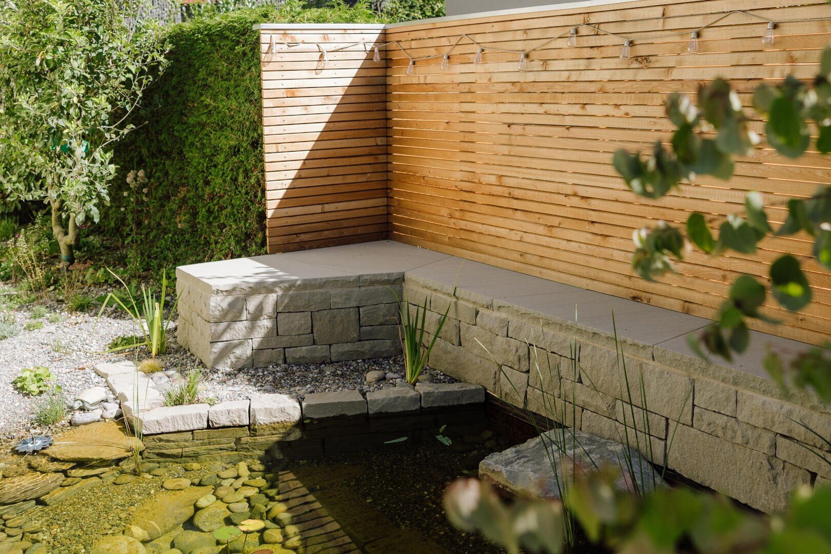 A small garden pond with clear water and stone edges lies next to a concrete and stone corner bench, behind which is a horizontal wooden fence with fairy lights. Green plants and shrubs surround the area.