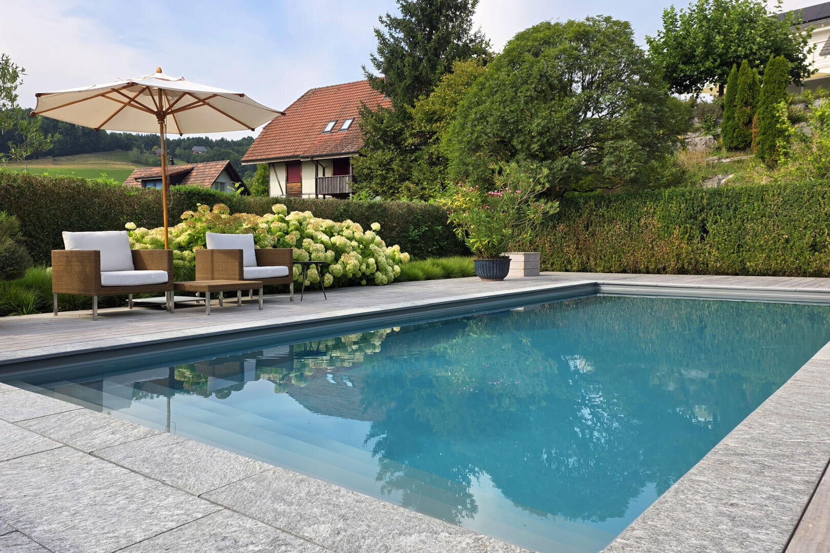 A modern backyard pool surrounded by stone tiles, with two wicker chairs and an umbrella on a wooden deck. Lush green bushes, trees, and a house with a red roof are in the background.