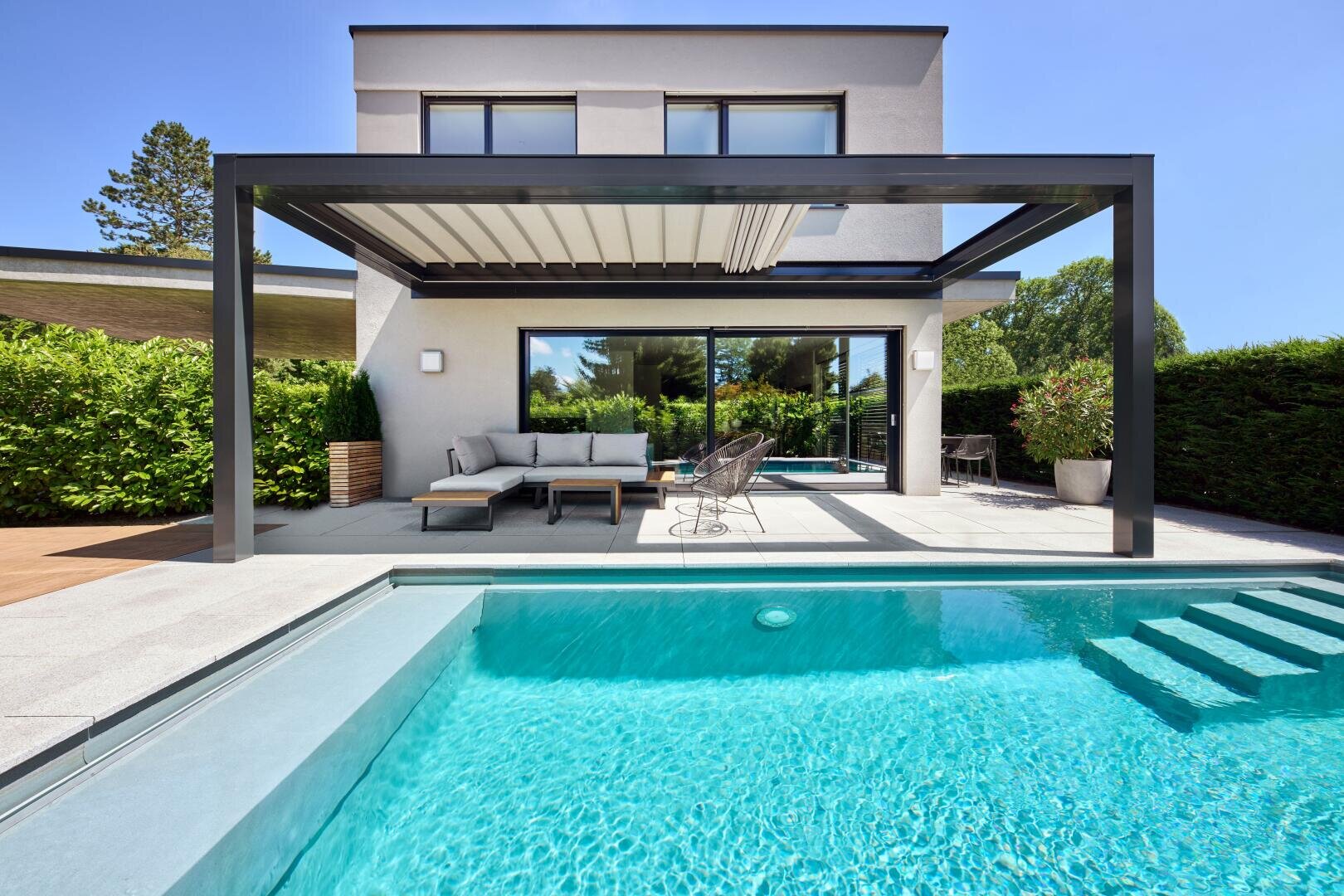 Modern house with a pergola-covered terrace, outdoor seating and a clear blue swimming pool in the foreground, surrounded by greenery and trees under a bright, sunny sky.