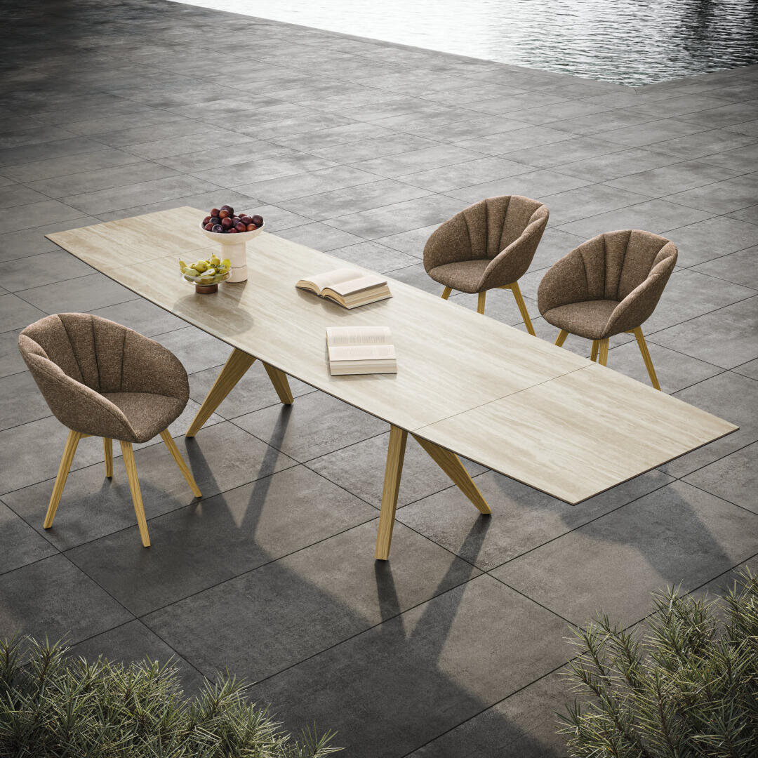 A modern dining table made of light wood with books, a bowl of apples and a small potted plant, surrounded by four brown upholstered chairs on a tiled terrace near a pool.