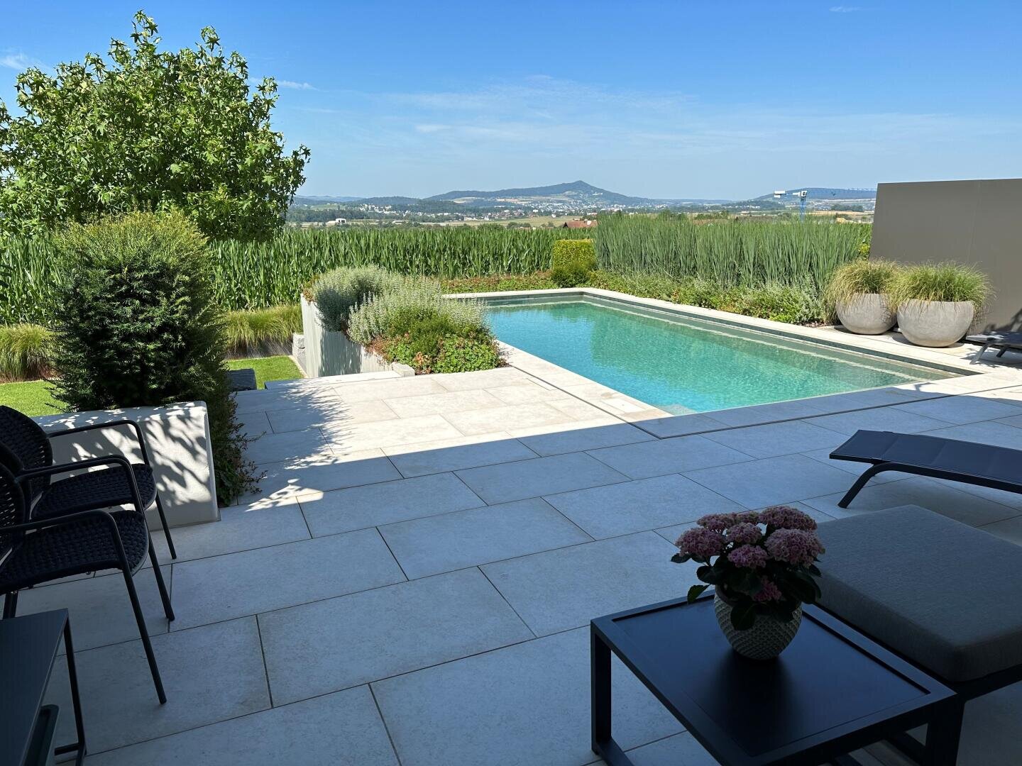 Ein moderner Garten mit einer Terrasse, Liegestühlen, Topfblumen und einem rechteckigen Swimmingpool, umgeben von Grün, mit Blick auf Felder und entfernte Hügel unter einem klaren blauen Himmel.