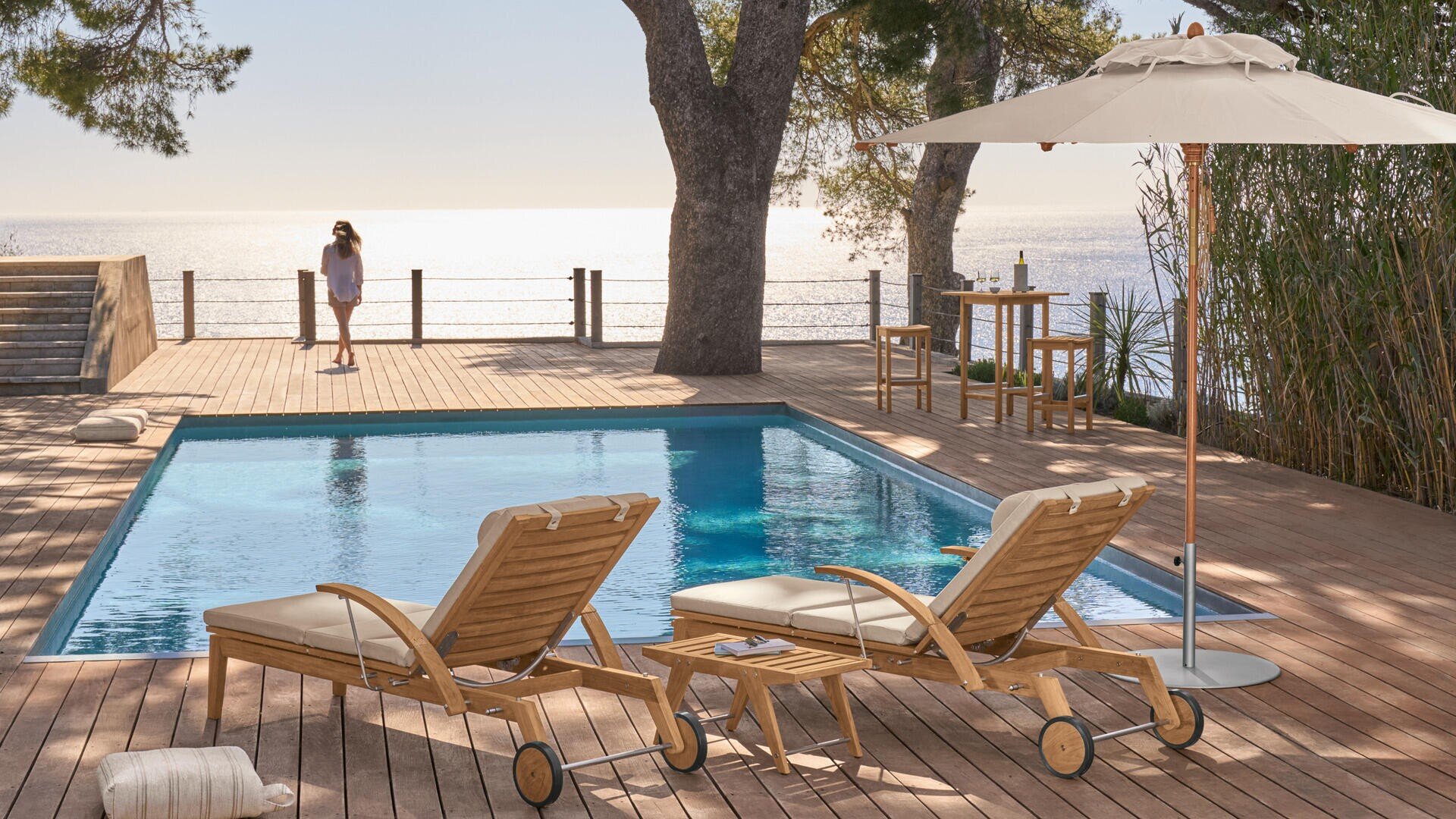 Zwei hölzerne Liegestühle mit Kissen stehen an einem Pool auf einem Holzdeck mit Blick auf das Meer. Im Hintergrund geht eine Frau spazieren. Ein Sonnenschirm und ein kleiner Tisch mit Stühlen befinden sich ebenfalls auf der Terrasse, umgeben von Bäumen und Pflanzen.