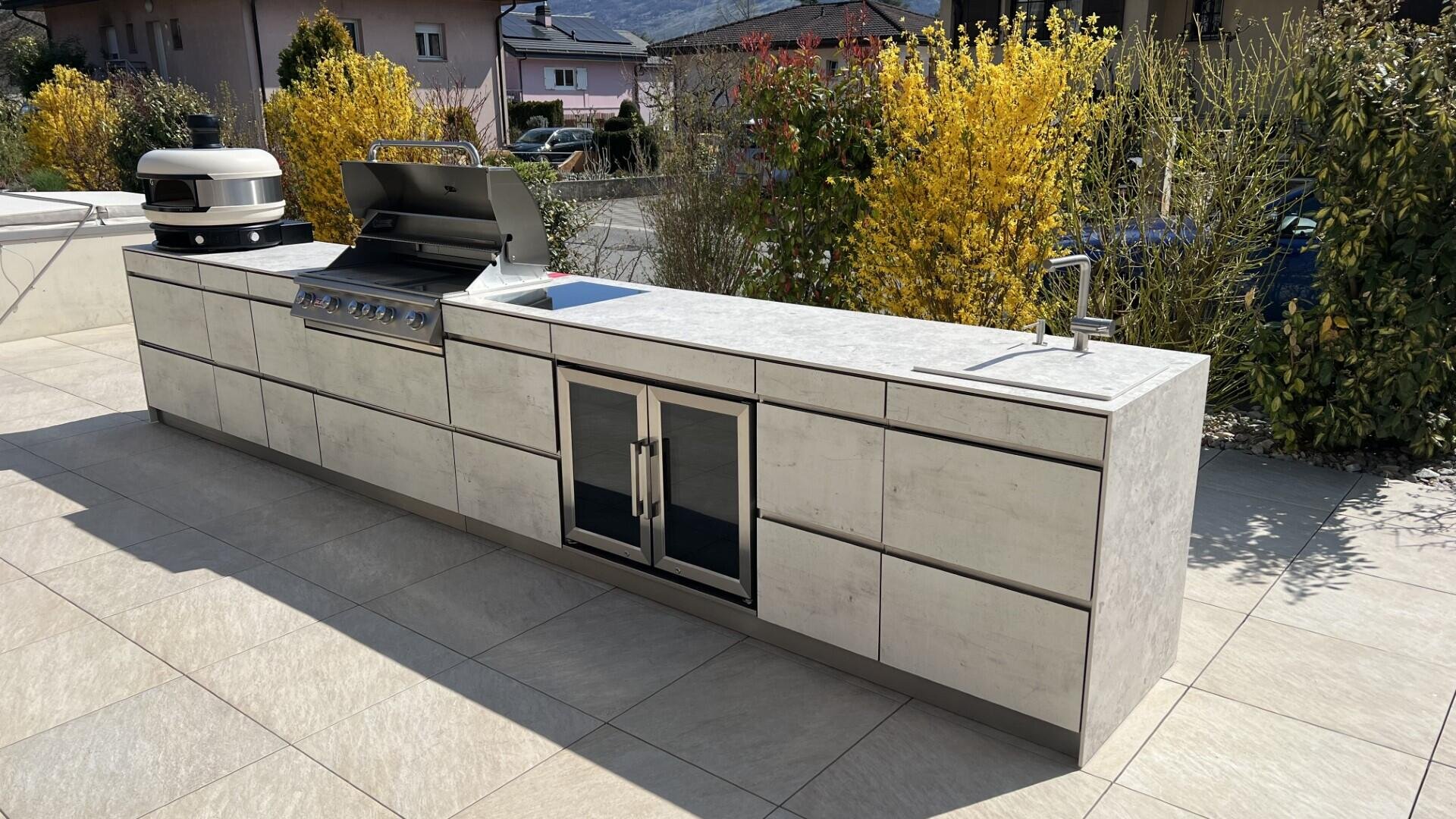 Moderne Außenküche mit eingebautem Grill, Pizzaofen, Spüle, Minikühlschrank und Schubladen auf einer gefliesten Terrasse mit gelb blühenden Sträuchern und Häusern im Hintergrund.