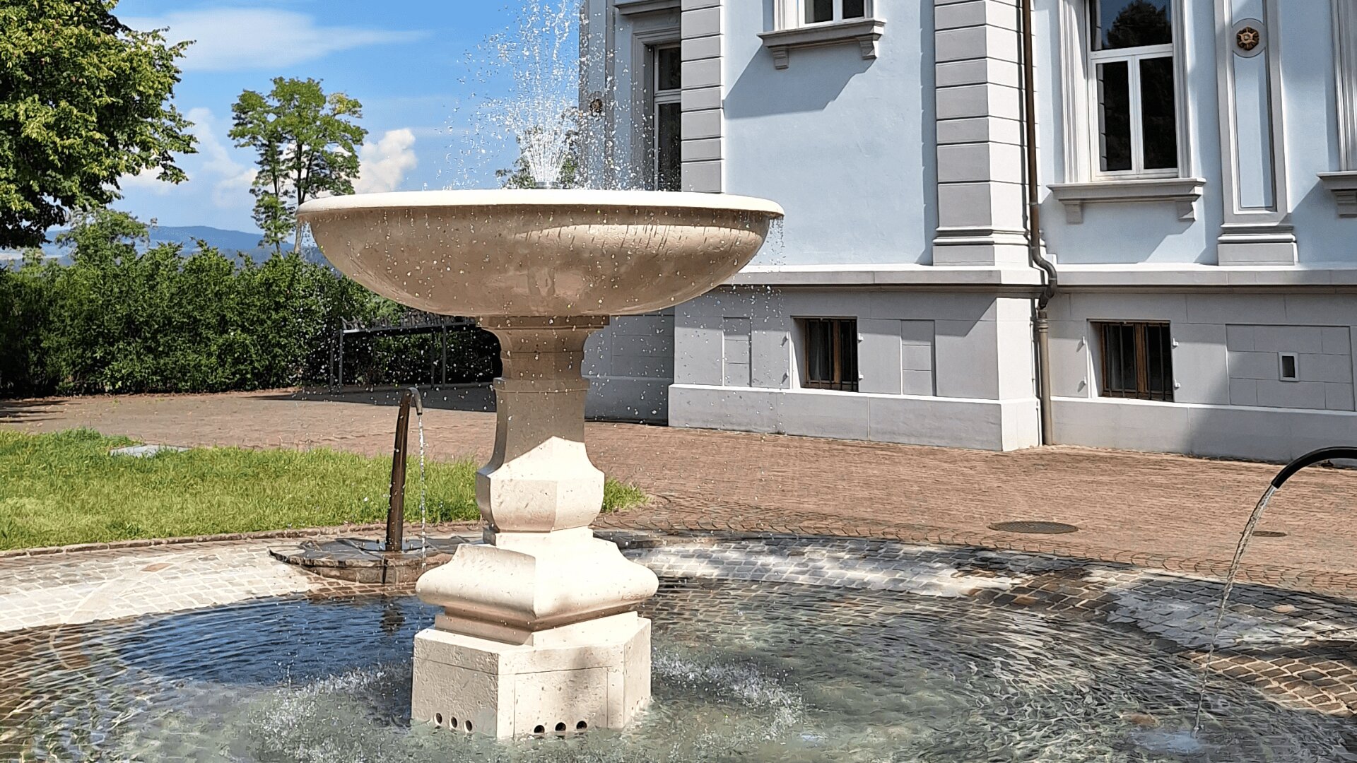 Vor einem hellgrauen, historischen Gebäude mit großen Fenstern und Zierleisten steht ein steinerner Springbrunnen, dessen Wasser in einem runden Becken nach oben spritzt. Im Hintergrund sind grüne Bäume und blauer Himmel zu sehen.