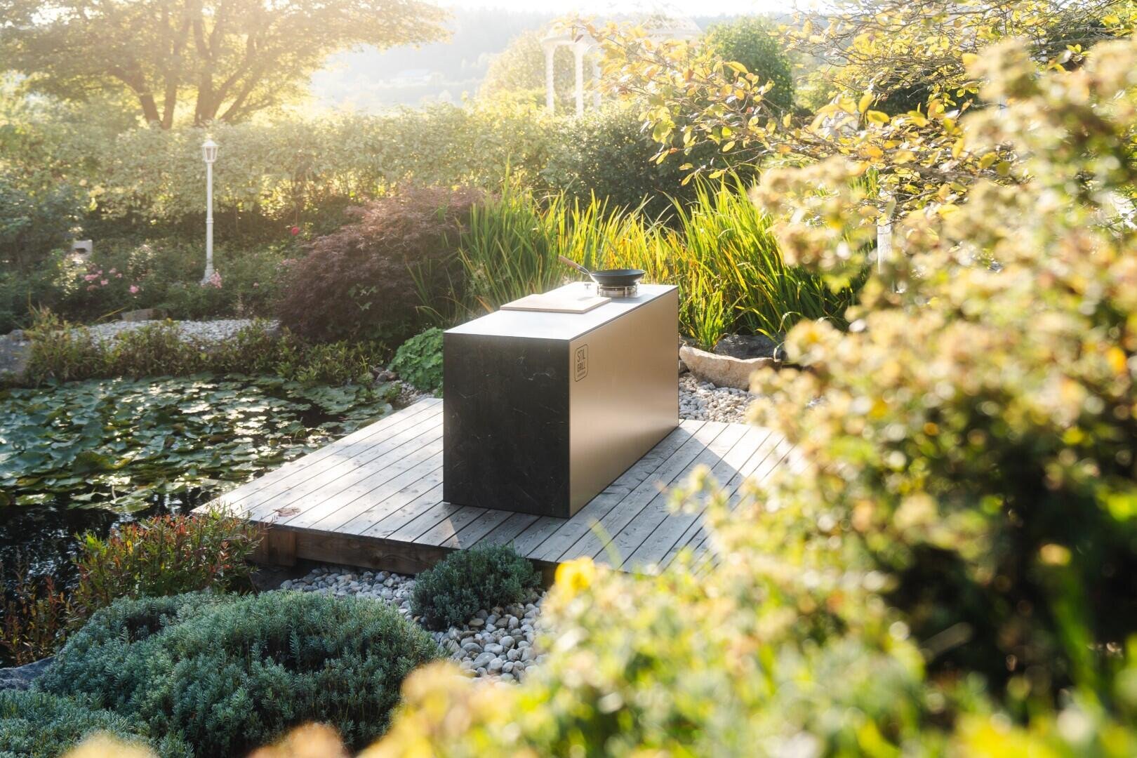 Ein moderner rechteckiger Außengrill steht auf einer Holzterrasse neben einem üppigen Gartenteich, umgeben von Grün und Pflanzen in sanftem Sonnenlicht.