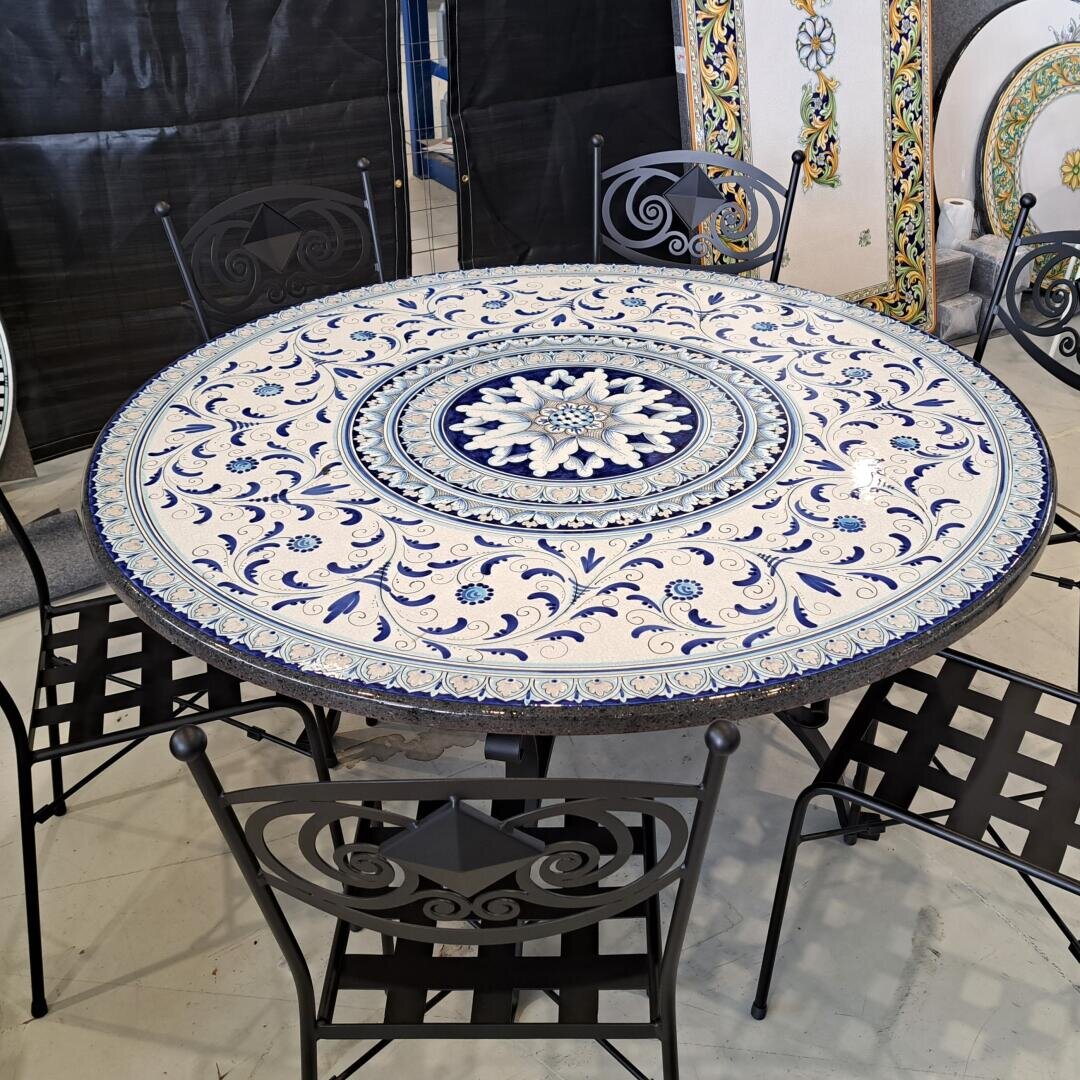 Ein runder Tisch mit einem detaillierten blau-weißen Mosaikmuster wird von vier schwarzen schmiedeeisernen Stühlen mit dekorativen Rückenlehnen und Sitzflächen umgeben, die in einem Innenraum auf einem hellen Boden stehen.