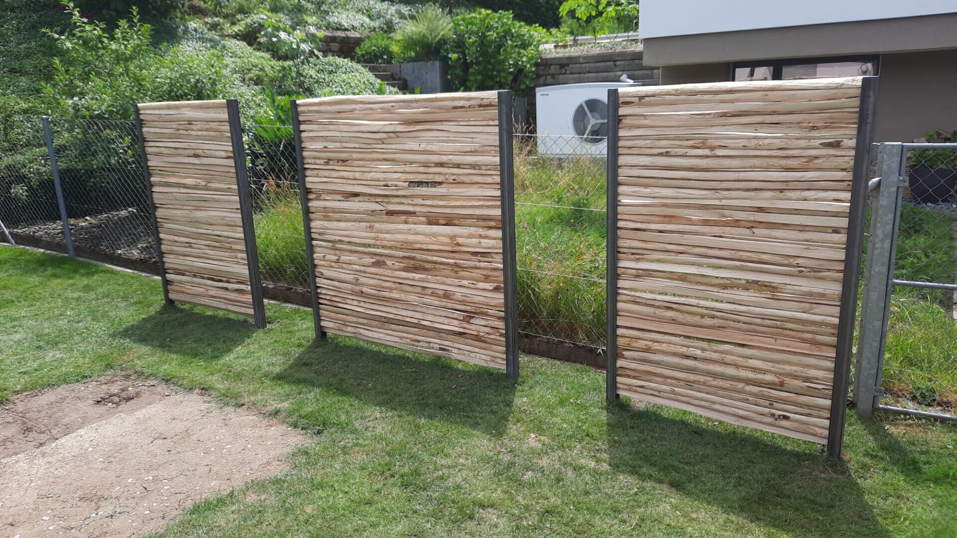 Drei hölzerne Sichtschutzwände mit horizontalen Lamellen stehen auf einem grasbewachsenen Rasen, der von Metallpfosten gestützt wird. Dahinter befinden sich ein Metallzaun, Grünflächen und ein Teil eines modernen Gebäudes mit einer Klimaanlage.