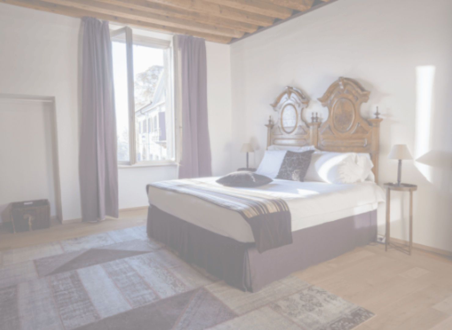 Ein helles Schlafzimmer mit einer Holzdecke, einem großen Fenster mit lila Vorhängen, einem Doppelbett mit verziertem Holzkopfteil, Beistelltischen mit Lampen und gemusterten Teppichen auf einem hellen Holzboden.