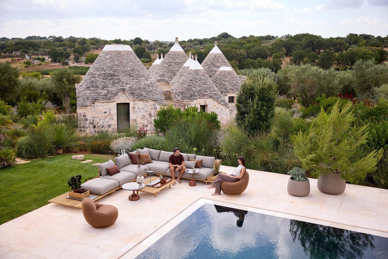 Zwei Personen entspannen sich auf modernen Außenmöbeln neben einem Pool, umgeben von üppigem Grün. Im Hintergrund stehen traditionelle Trulli-Häuser aus Stein mit kegelförmigen Dächern, eingebettet in eine ländliche Landschaft unter einem teilweise bewölkten Himmel.