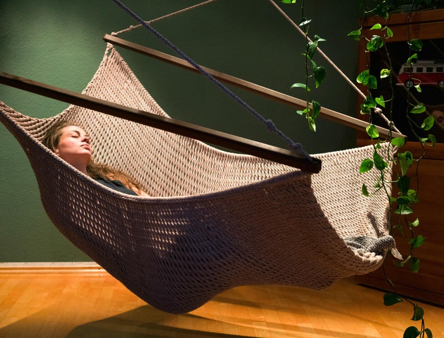 Eine Frau mit langen Haaren ruht friedlich in einer beigefarbenen, geflochtenen Hängematte in einem Haus, mit grünen Pflanzen in der Nähe und warmem Sonnenlicht, das den Holzboden beleuchtet.
