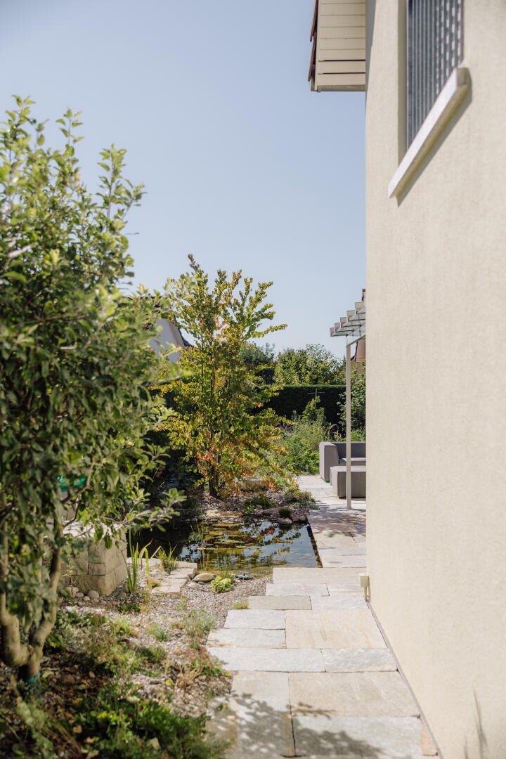 A stone path leads past a beige house and a small pond surrounded by greenery and trees under a clear blue sky.