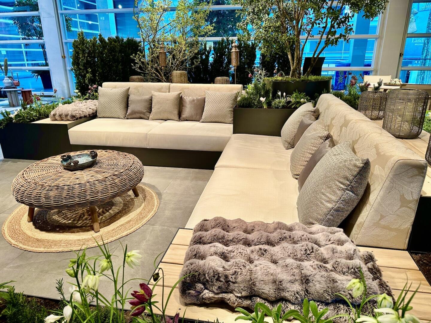 Moderner Lounge-Bereich im Freien mit zwei beigen Sofas, Plüschkissen, einem runden Couchtisch aus Weidengeflecht und Fellüberwürfen. Umgeben von Pflanzen und Grün, strahlt die Umgebung eine gemütliche, einladende Atmosphäre aus.