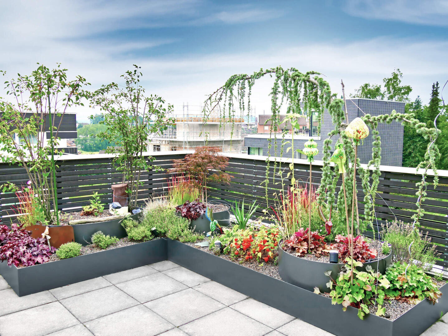 Ein moderner Dachgarten mit verschiedenen grünen und blattreichen Pflanzen in Hochbeeten, umgeben von einem dunklen Holzzaun, mit Gebäuden und einem bewölkten Himmel im Hintergrund.