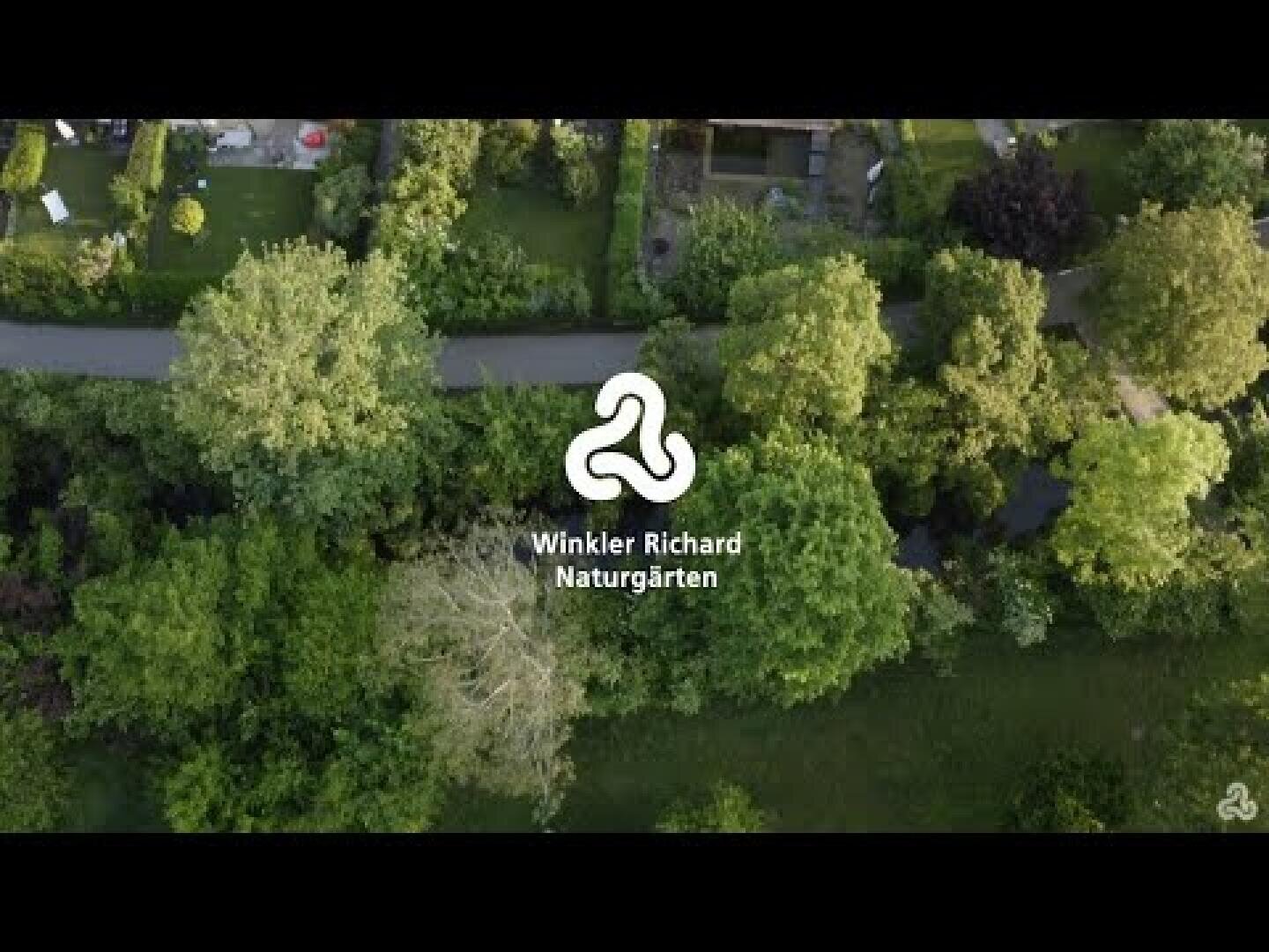 Luftaufnahme eines üppigen, grünen Gartens mit verschiedenen Bäumen und Sträuchern. In der Mitte ein weißes Logo und die Aufschrift Winkler Richard Naturgärten. Eine Straße ist sichtbar, die die Gartenbereiche trennt.