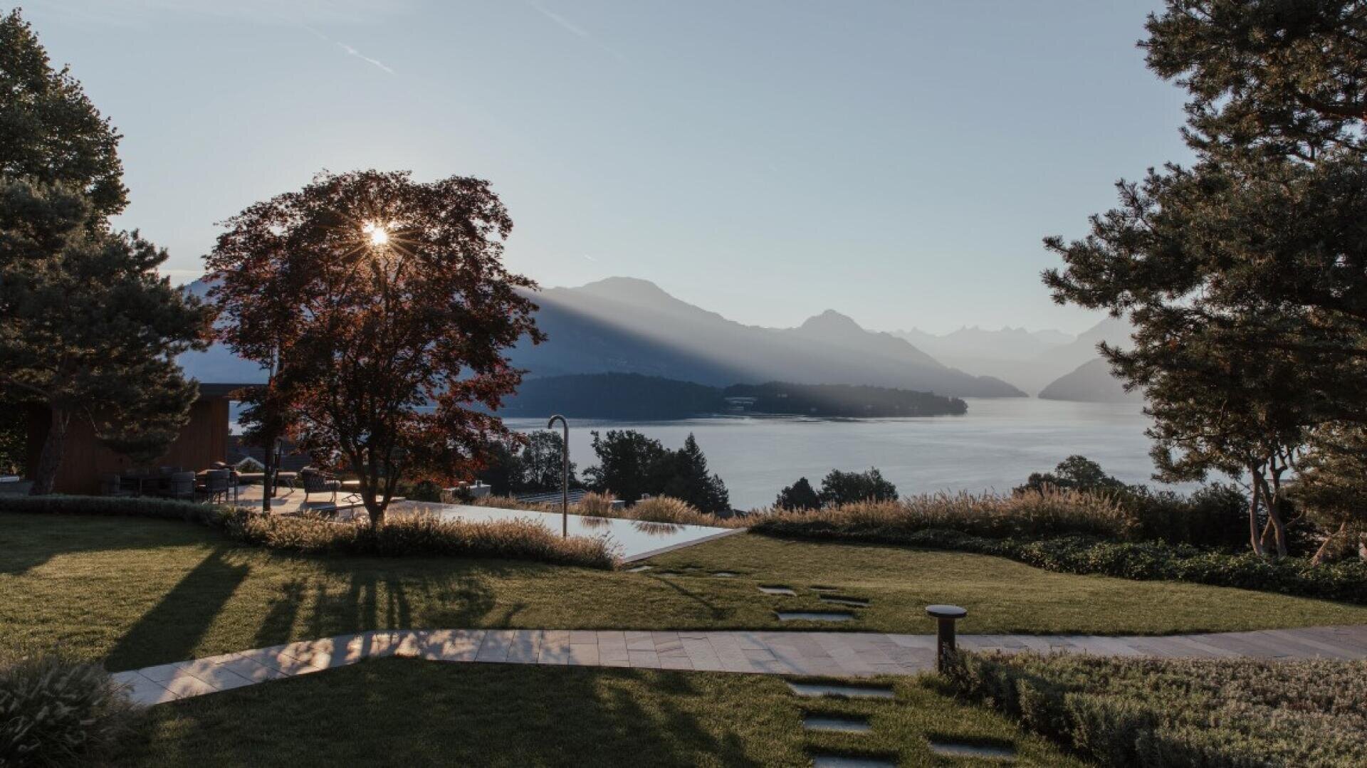 Eine heitere Aussicht auf einen See bei Sonnenaufgang. Das Sonnenlicht fällt durch einen Baum und wirft lange Schatten auf einen grasbewachsenen Garten. In der Ferne sind Berge und Wasser unter einem klaren Himmel zu sehen.
