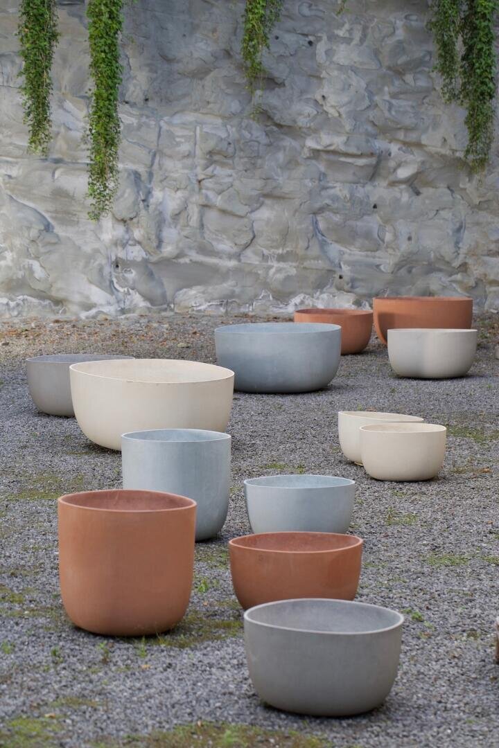 Große, leere Pflanztöpfe in Terrakotta-, Beige- und Grautönen stehen auf Kies vor einer Steinmauer, von der grüne Ranken herabhängen.