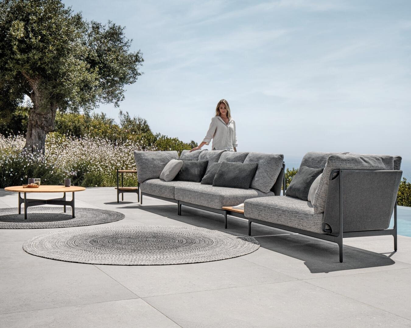Eine Frau steht neben einem modernen grauen Outdoor-Sectional-Sofa mit Kissen auf einer Steinterrasse in der Nähe eines Pools. Ein runder Teppich und ein hölzerner Couchtisch sind in der Nähe angeordnet. Grüne Bäume und Sträucher umgeben den Bereich unter blauem Himmel.