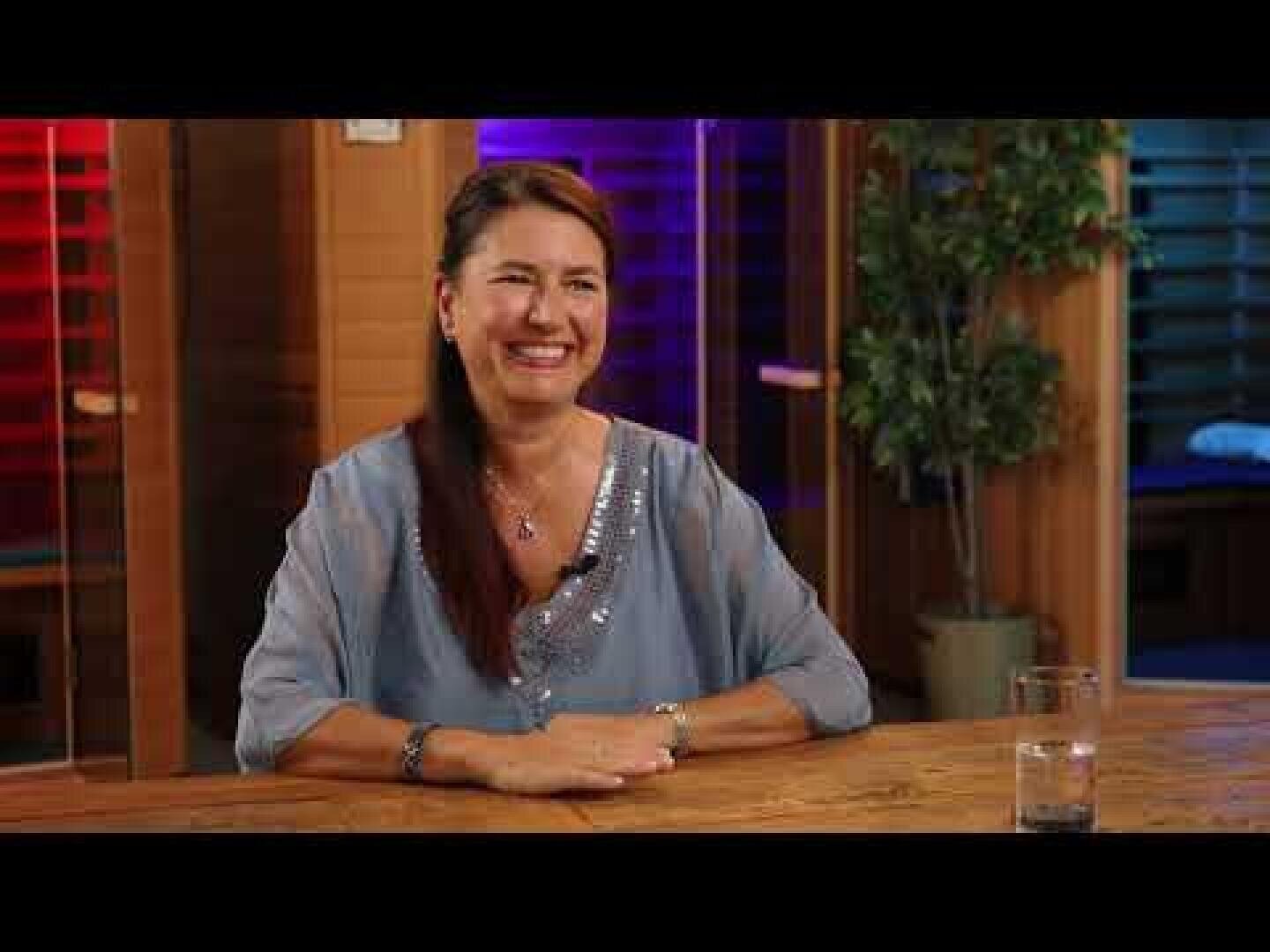 A woman with long brown hair and a light blue blouse sits smiling at a wooden table inside the room. There is a glass of water on the table and in the background are colored, blurry lights and a potted plant.