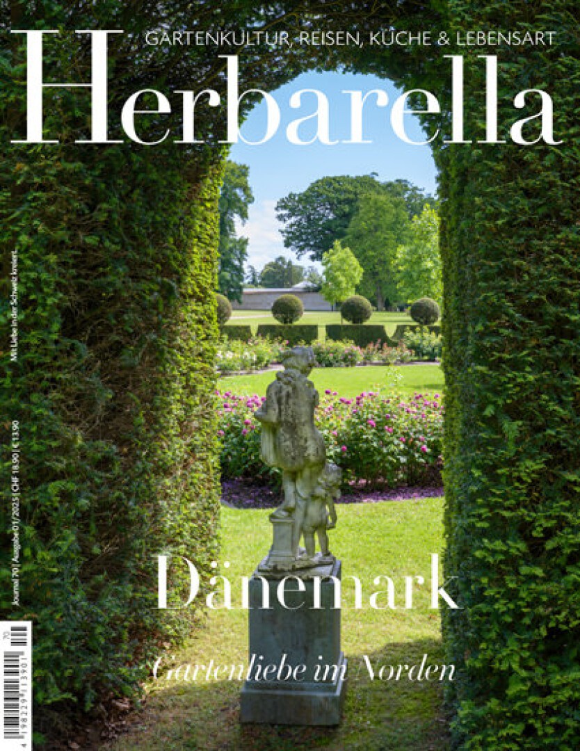 Eine Gartenszene, die durch eine hohe, bogenförmige Heckenöffnung betrachtet wird. Im Vordergrund steht eine klassische Statue, im Hintergrund sind gepflegte Rasenflächen und Bäume zu sehen. Der Covertext lautet „Herbarella“ und enthält Informationen über Dänemark.