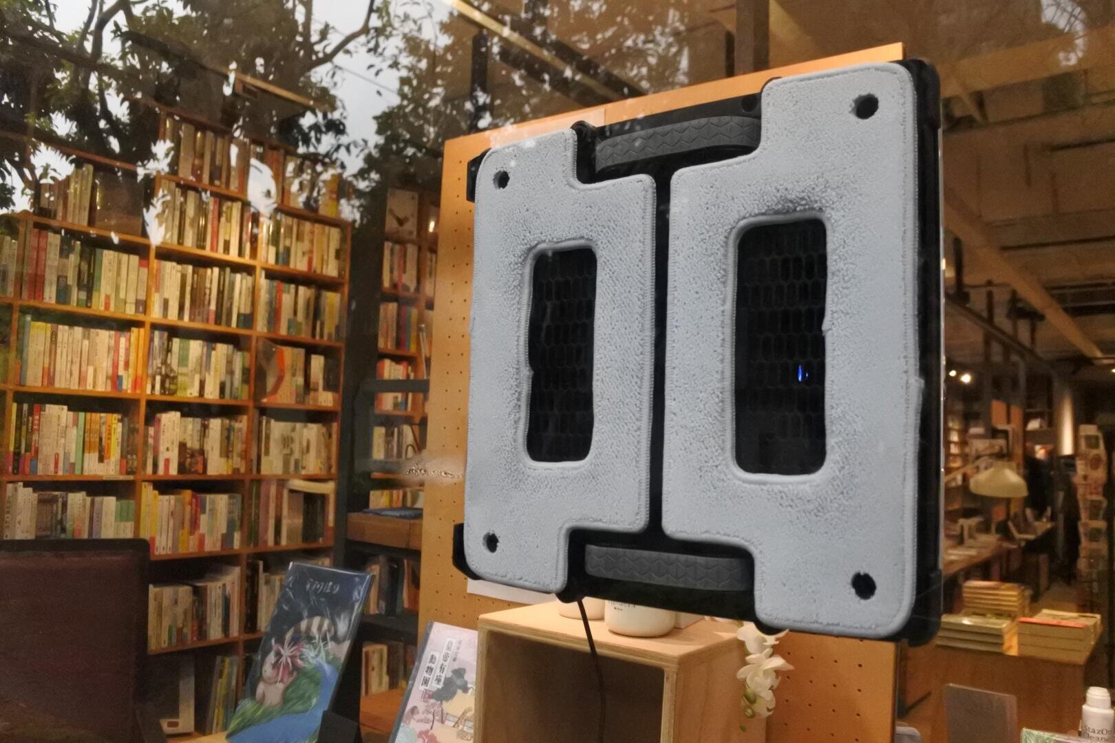 Ein Fensterreinigungsroboter ist an einem Glasfenster in einer gemütlichen Buchhandlung mit vielen Bücherregalen angebracht und erzeugt eine Spiegelung des Innenraums und der Bäume draußen.