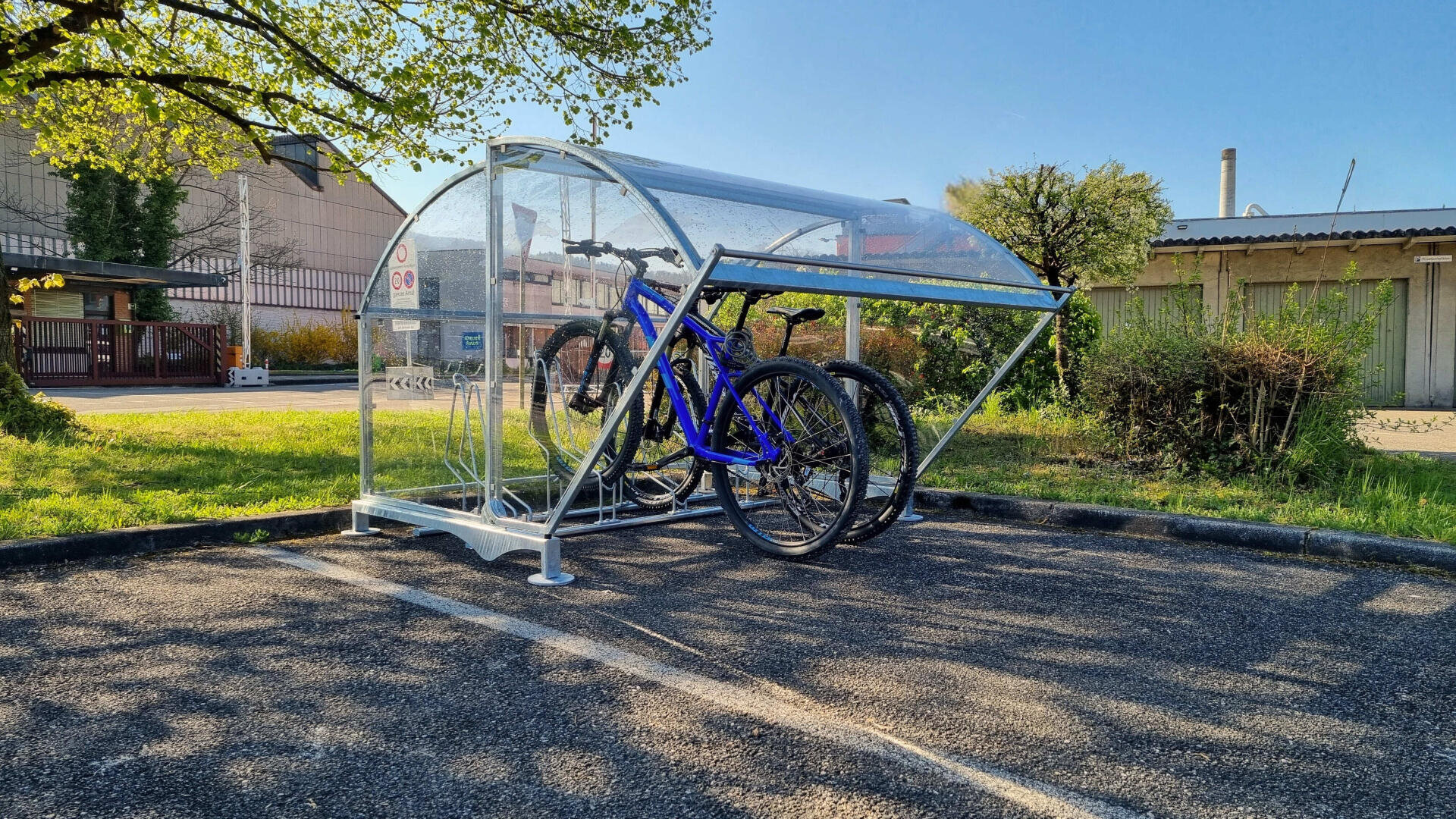 Ein transparenter Fahrradunterstand mit einem gewölbten Dach steht auf einem Parkplatz und beherbergt an einem sonnigen Tag drei Fahrräder, mit Bäumen und Gebäuden im Hintergrund.