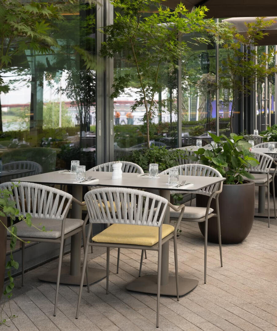 Außenbereich mit modernen Tischen und Stühlen, ausgestattet mit Gläsern und Besteck, umgeben von großen Topfpflanzen und Grünpflanzen, neben einem Gebäude mit Glaswänden.