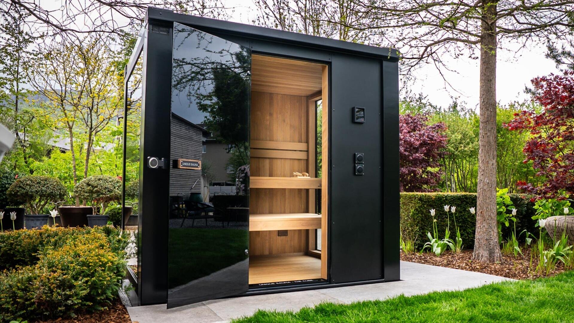 Eine moderne Außensauna mit schwarzem Äußeren und einer Glastür, die auf einer von Bäumen, Sträuchern und Gartenpflanzen umgebenen Steinterrasse steht. Der hölzerne Innenraum ist mit Sitzbänken und natürlichem Licht ausgestattet.
