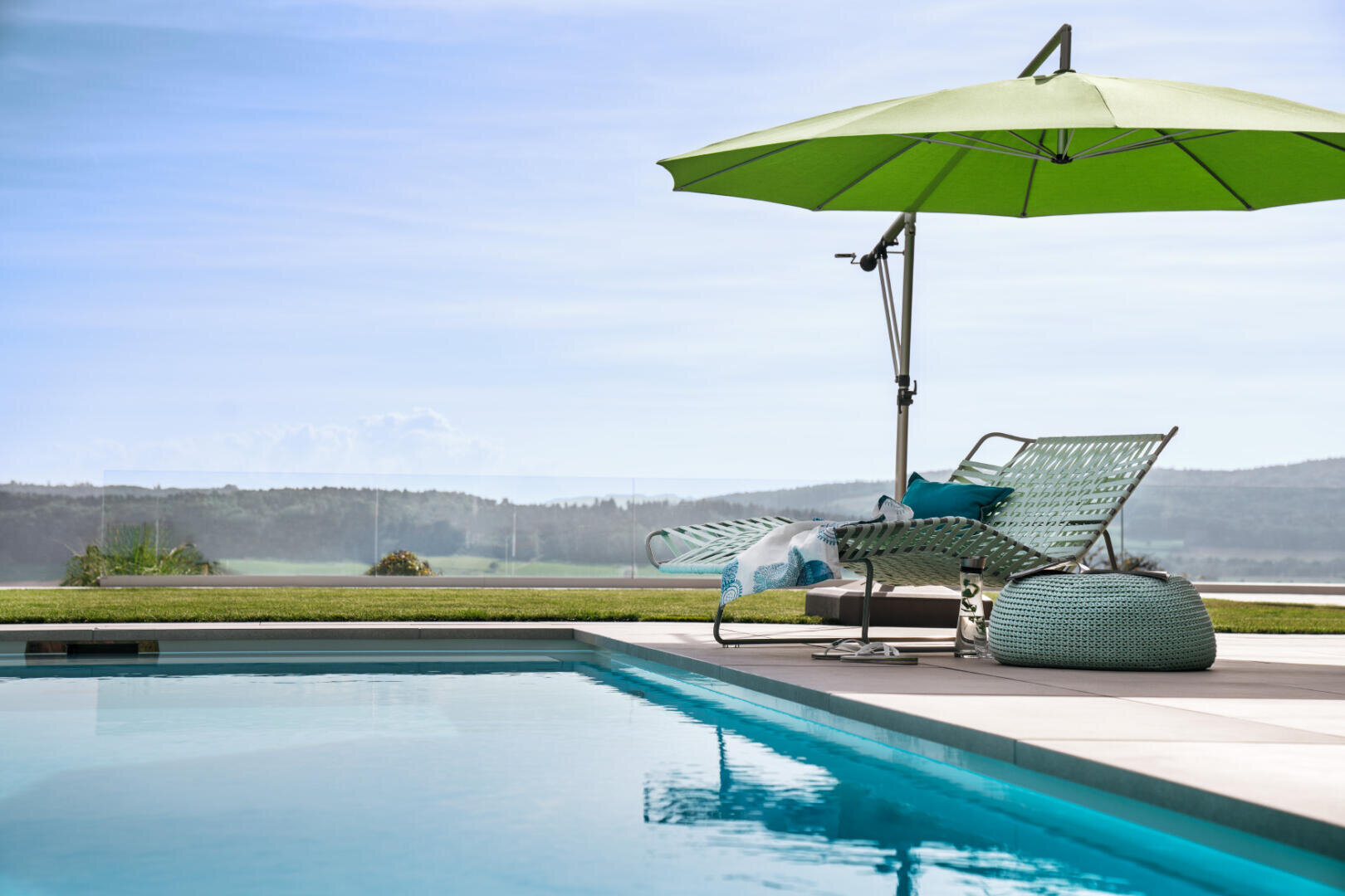Ein Loungebereich am Pool mit einem grünen Sonnenschirm, zwei geflochtenen Liegestühlen, einem kleinen Tisch und einem blaugrünen Hocker mit Blick auf grasbewachsene Felder und Hügel unter einem klaren blauen Himmel.