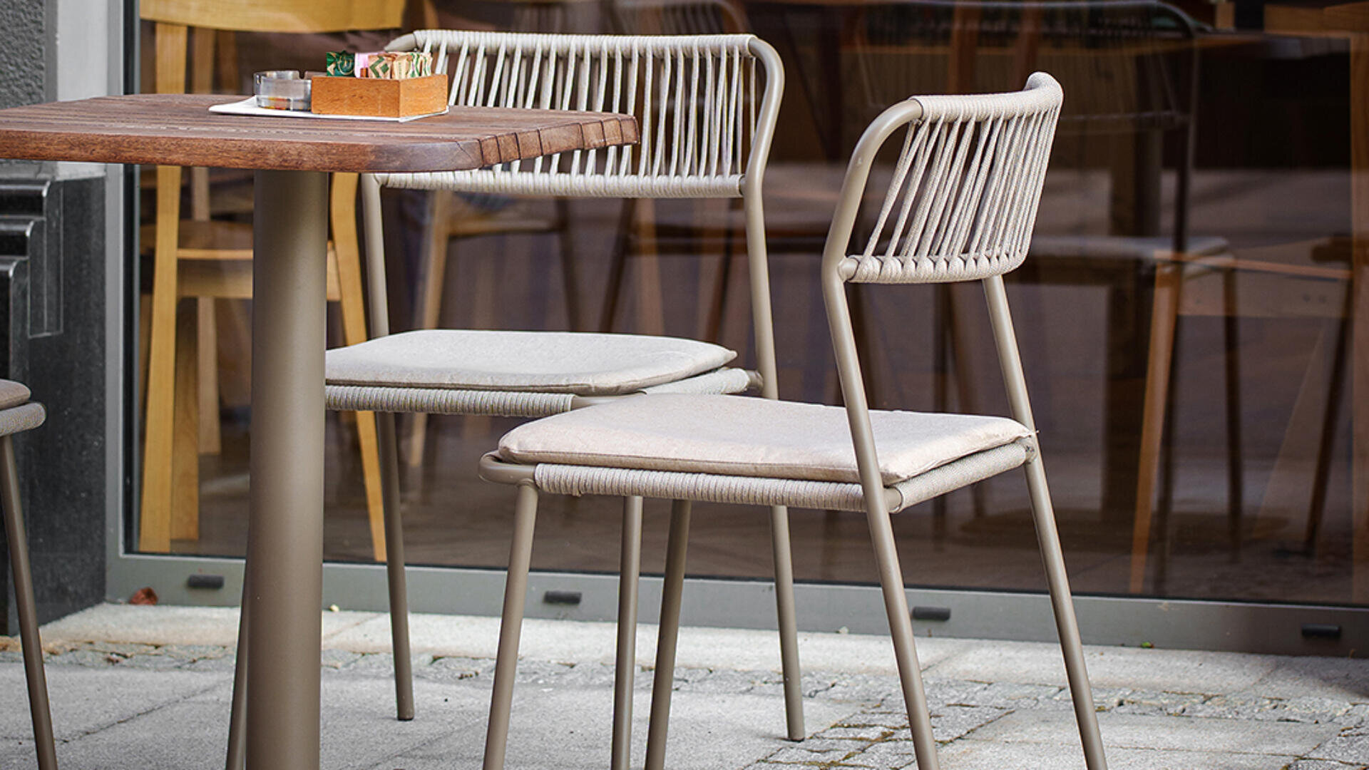 Zwei moderne Outdoor-Stühle mit hellen Kissen sitzen an einem kleinen Holztisch auf einer Terrasse, während ein weiterer Tisch und Stühle durch ein großes Fenster im Hintergrund zu sehen sind.