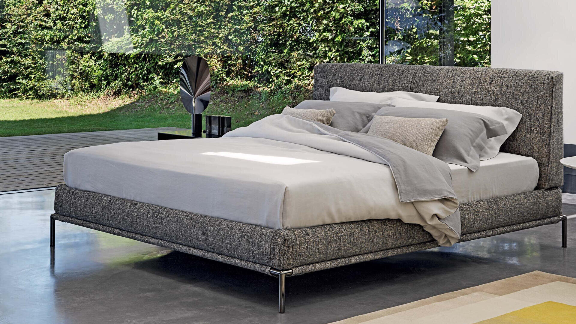 A modern bedroom with a gray upholstered bed, neatly covered with gray and beige bedding, stands on a polished floor near large windows overlooking a green garden. A decorative object and a small table stand next to the bed.